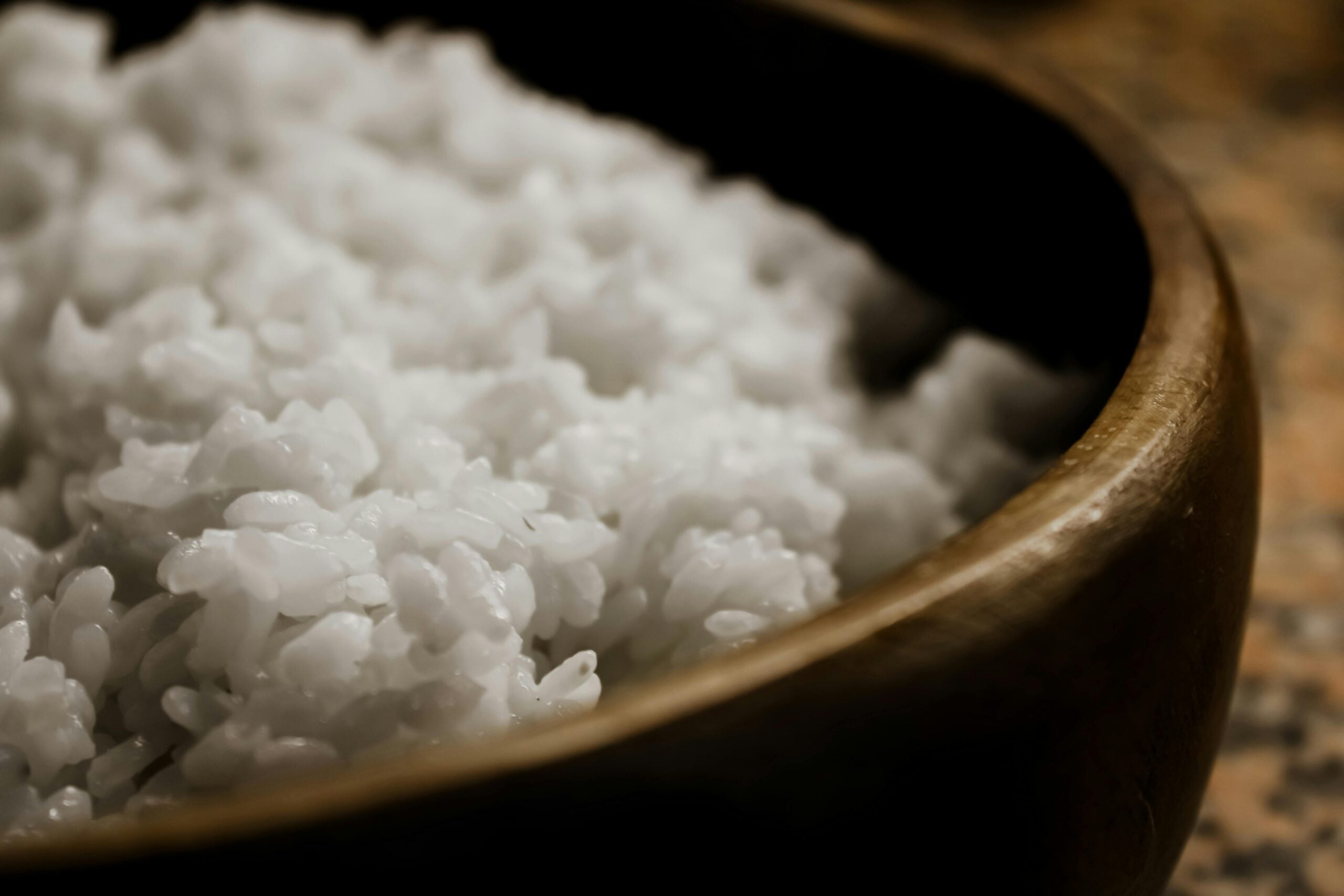 Close-up of steamed white rice in a wooden bowl, with rice grains appearing soft and slightly glossy. The background is blurred, drawing focus to the texture of the rice.