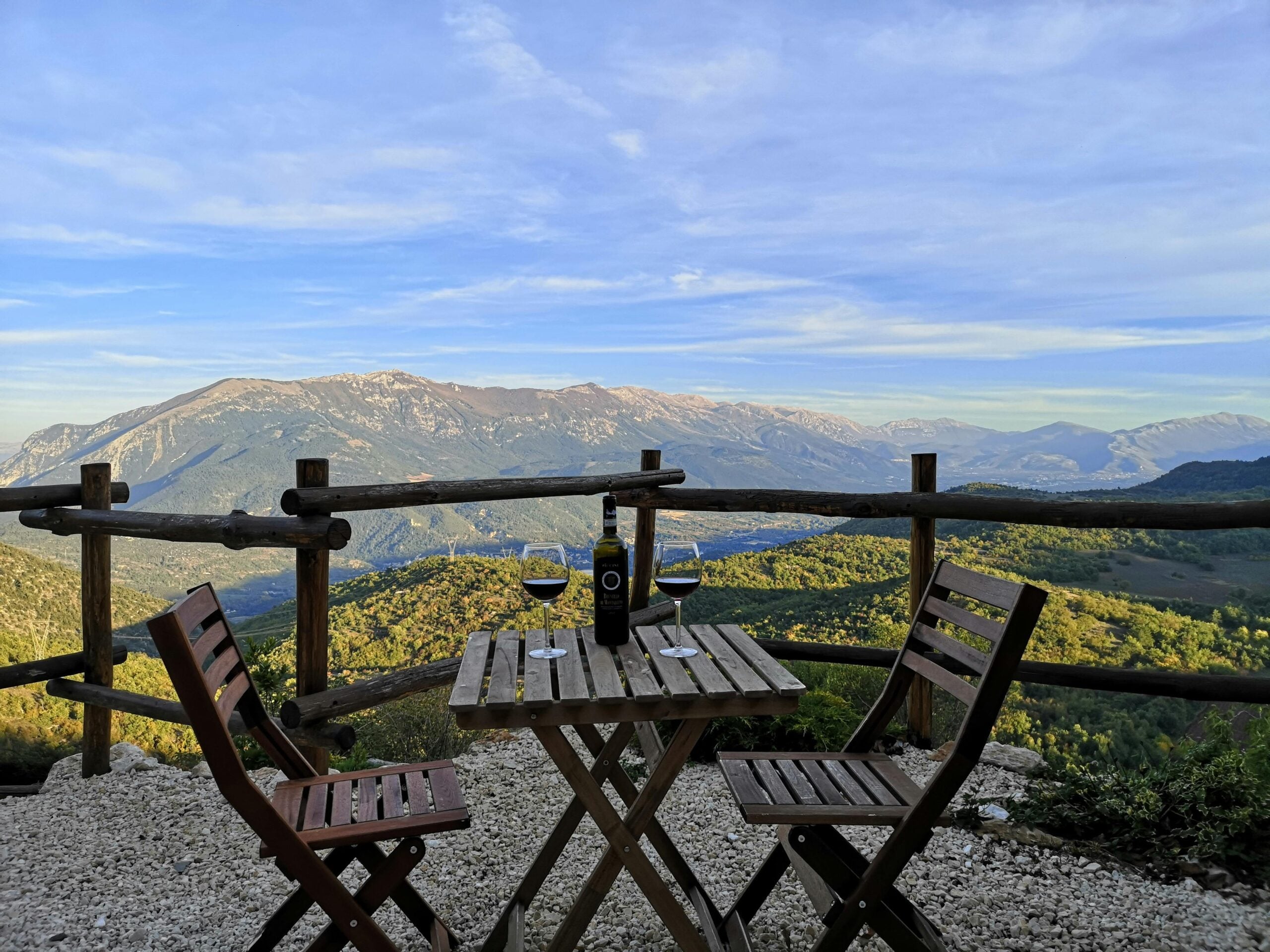A wooden table with two chairs overlooks a scenic mountain landscape. On the table are two glasses of red wine, a wine bottle, and plates, with a rustic wooden fence and blue sky in the background.
