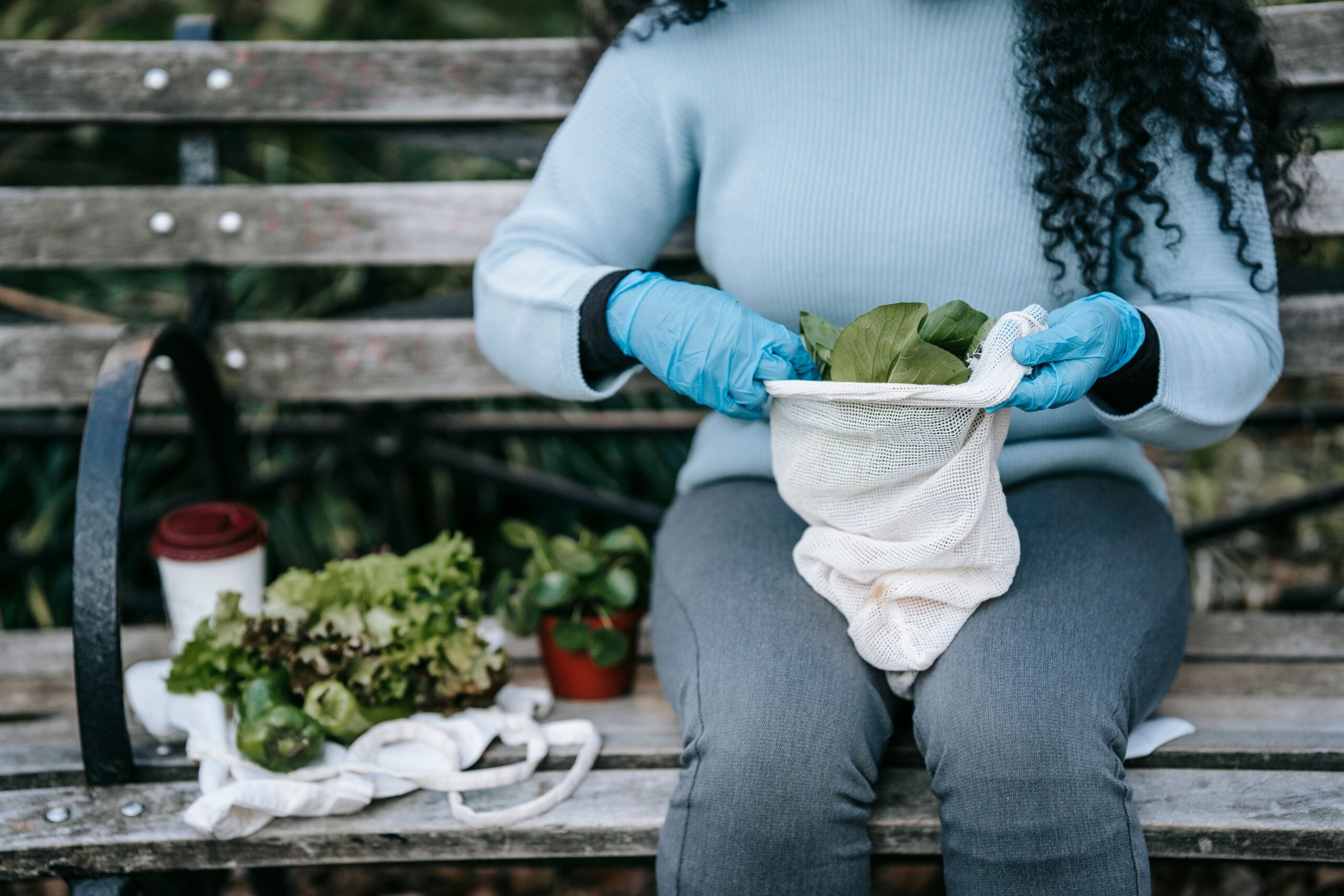 A person wearing blue gloves and a light blue sweater sits on a bench, holding a cloth bag with leafy greens. Fresh vegetables and a coffee cup rest beside them on the bench.