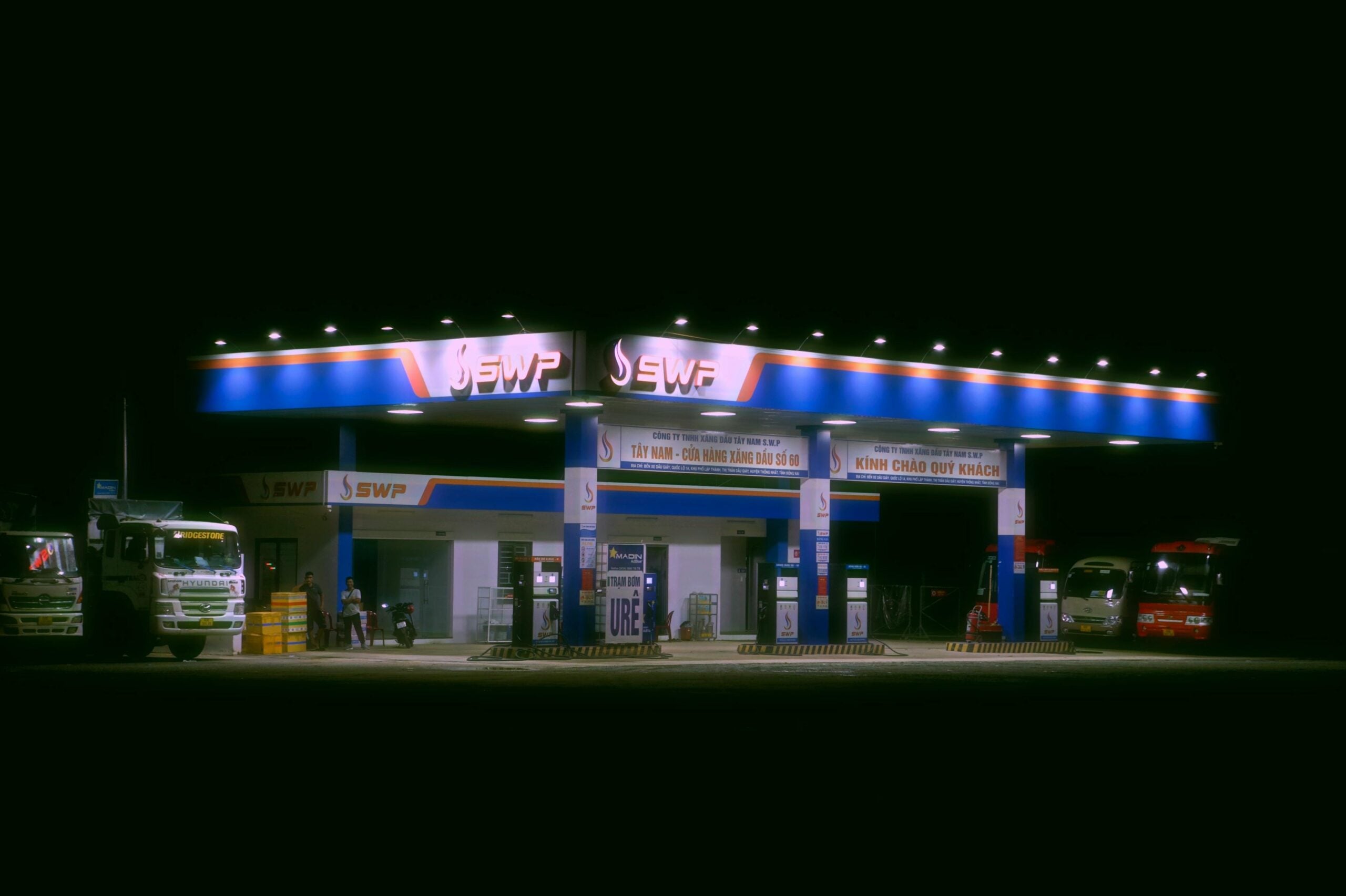 A brightly lit gas station at night with blue and orange signage. Several trucks are parked by the pumps, and a few people are visible. The surrounding area is dark, highlighting the station’s lights.
