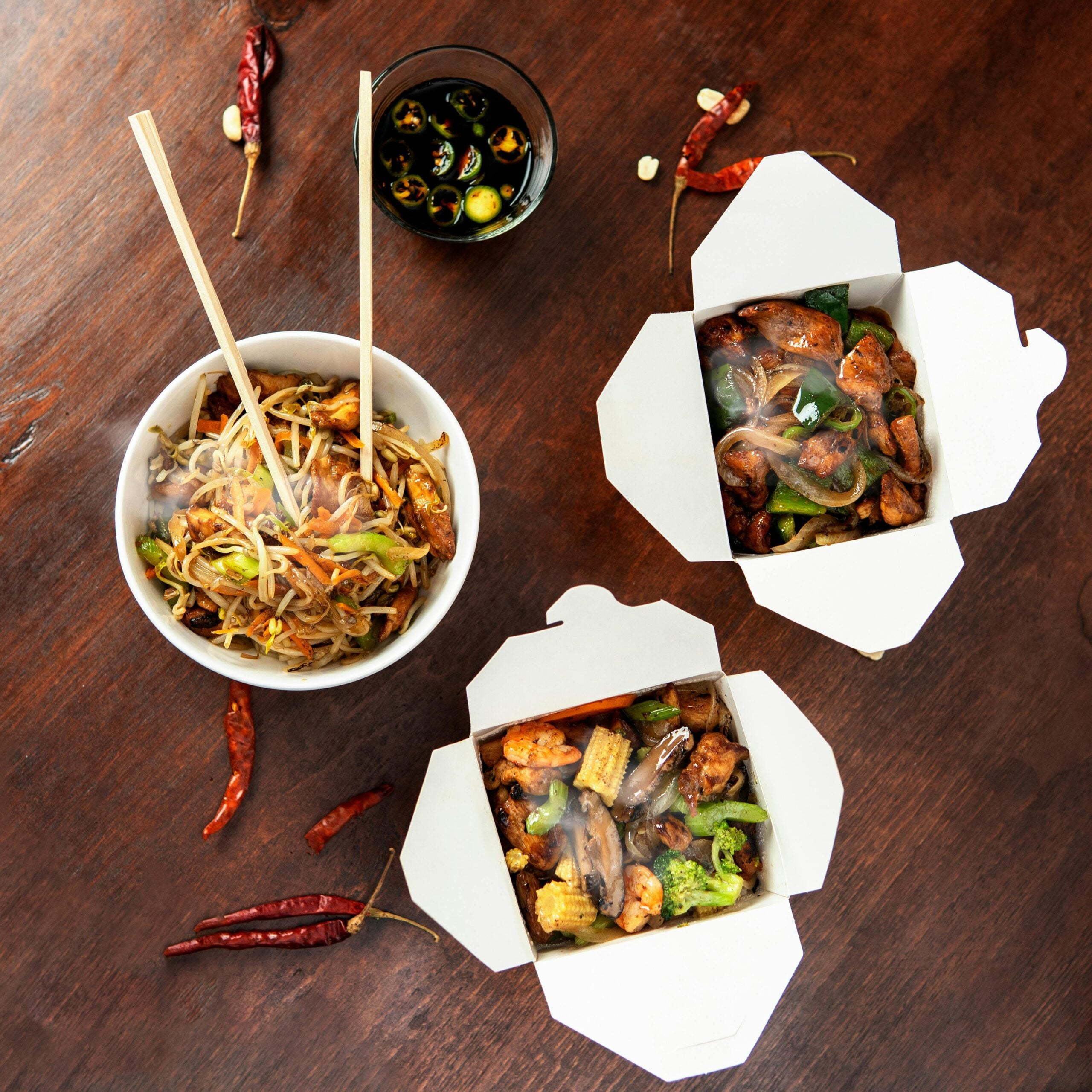 A bowl of stir-fried noodles with chopsticks, two open takeout boxes with vegetables and meat, a small bowl of sliced chilies in soy sauce, and dried red chilies on a wooden table.