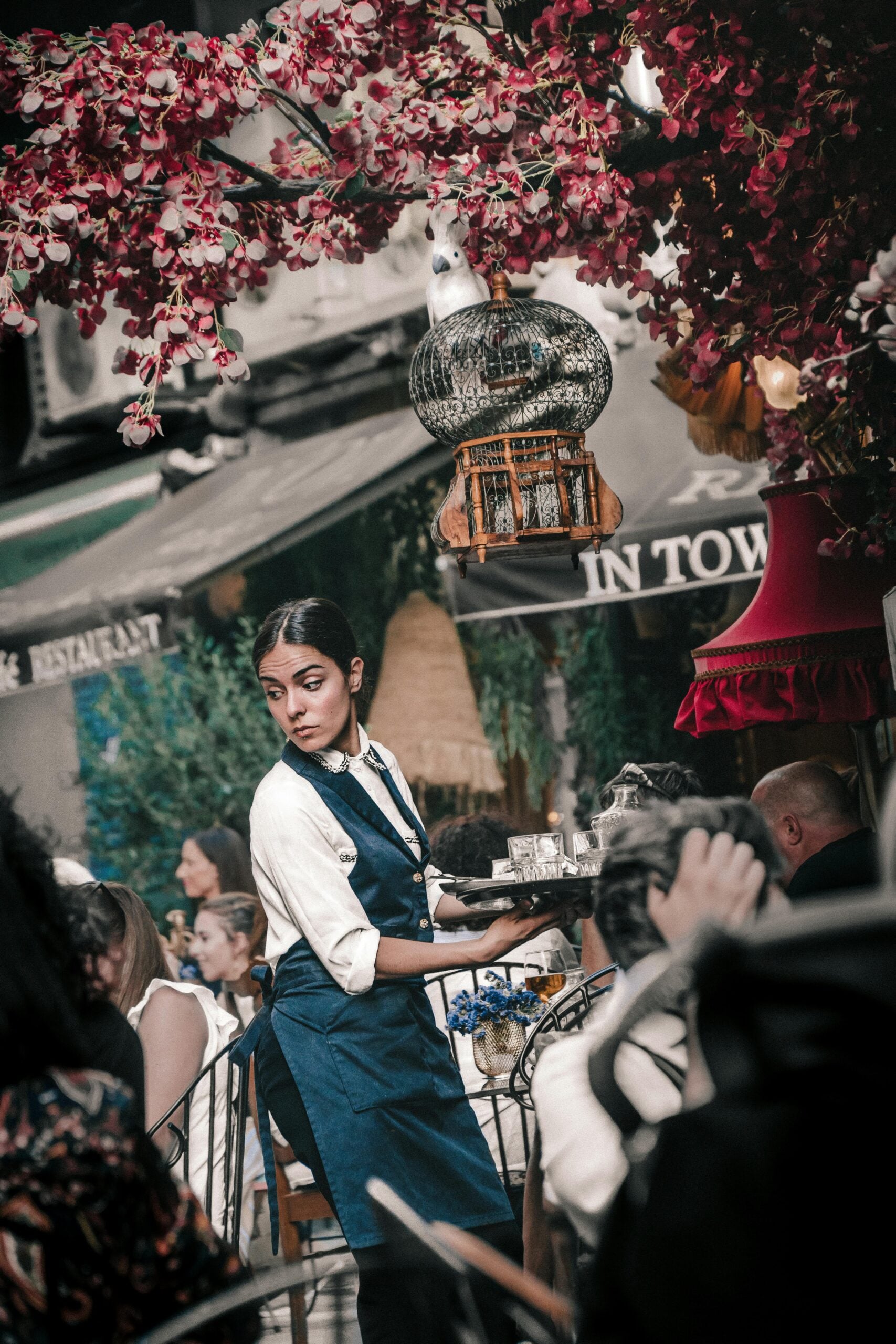 A waitress in a blue apron serves drinks at an outdoor café filled with people. Pink flowers and a decorative birdcage hang overhead, creating a lively and vibrant atmosphere.