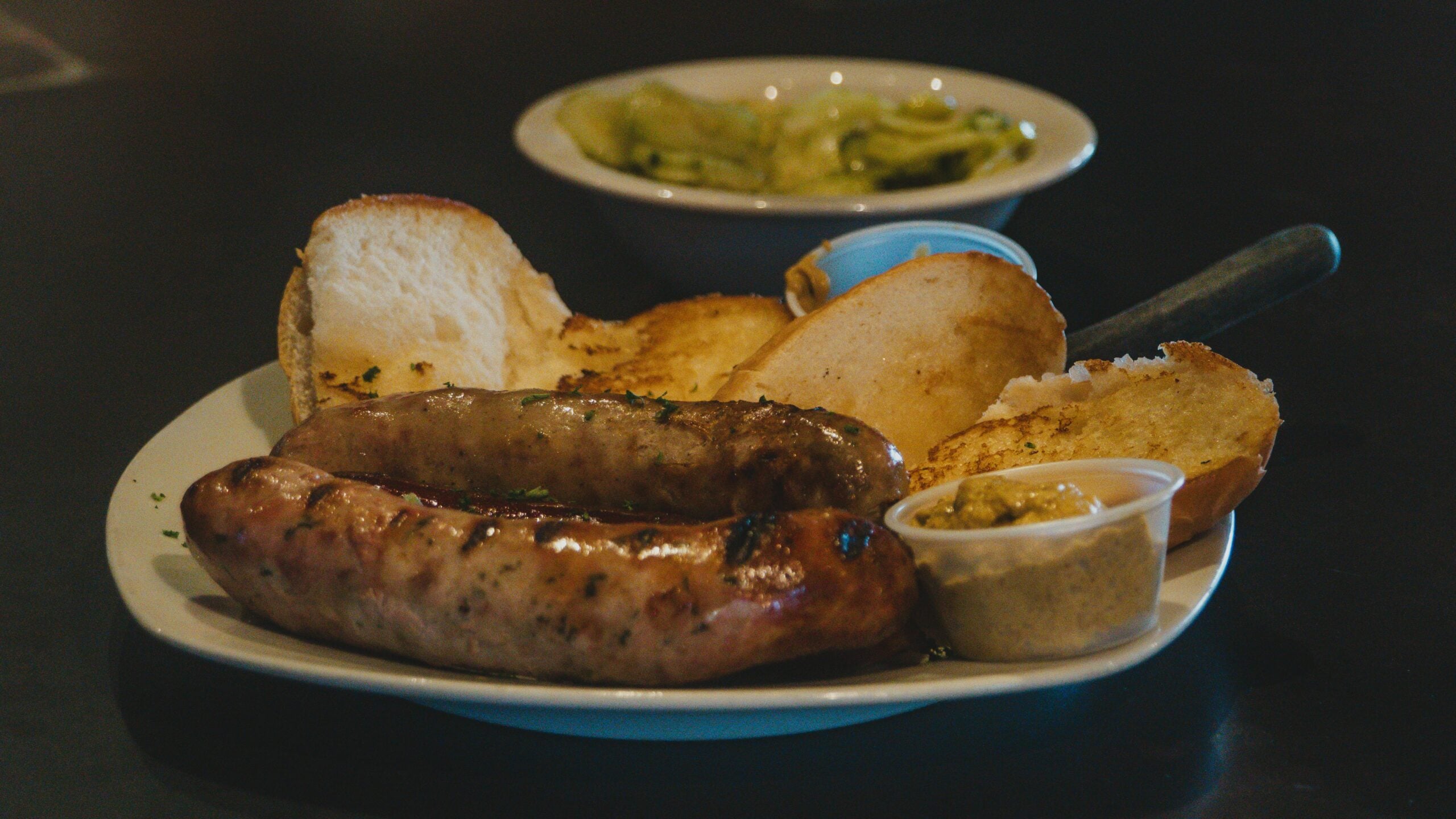 A plate with two grilled sausages, toasted bread slices, and a small container of mustard sits in the foreground, with a bowl of sliced cucumbers in the background.