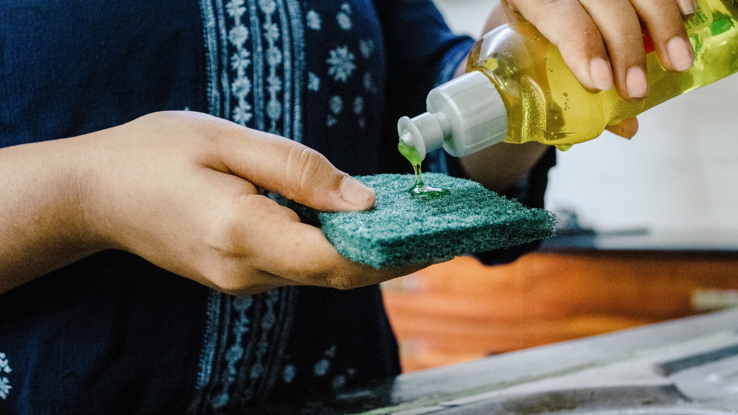 A person pours green dishwashing liquid onto a green scrub sponge while standing by a kitchen sink. The person is wearing a navy blue top with white embroidery.