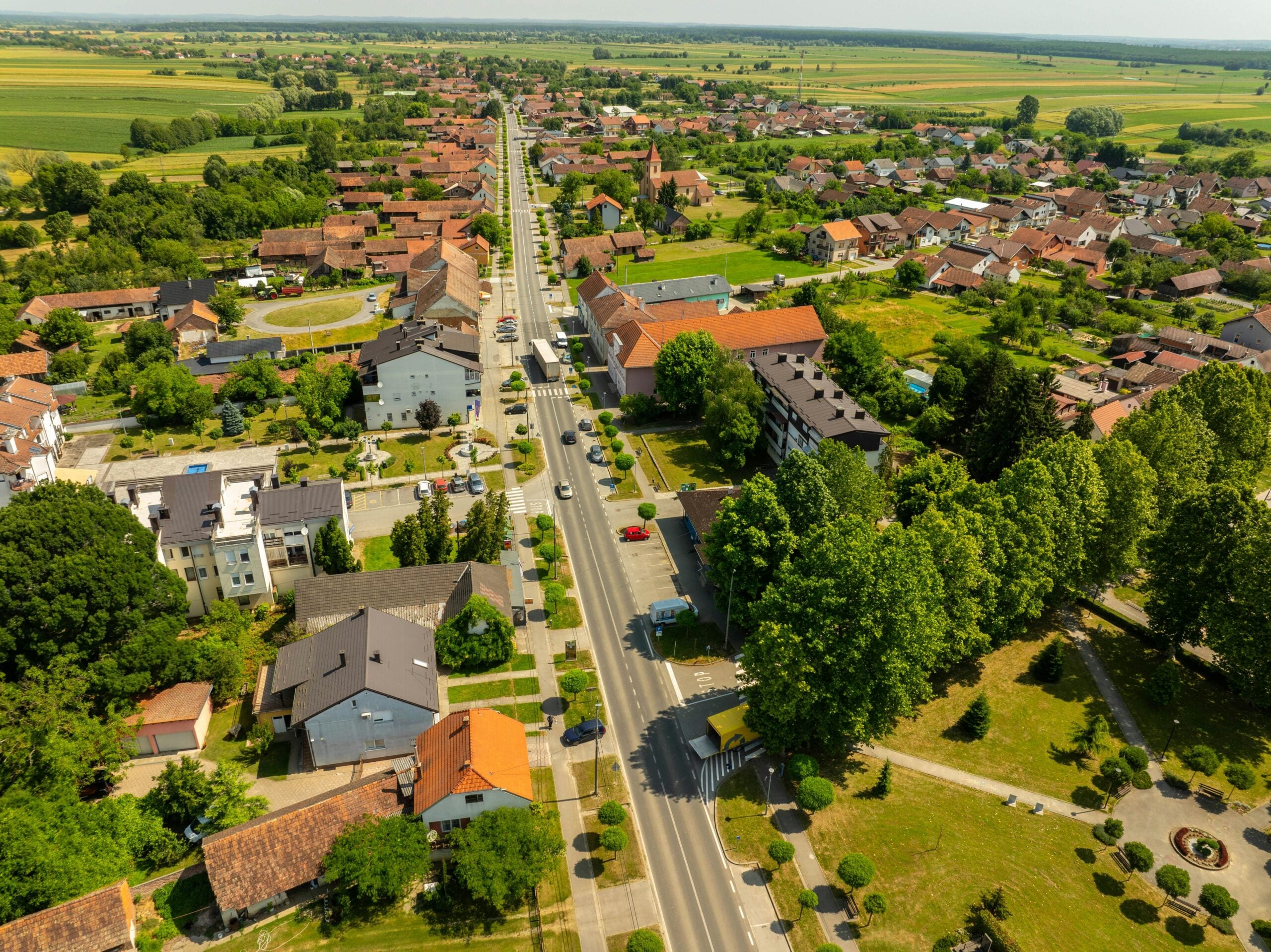 Aerial view of a small town with a main road running through the center, surrounded by houses, buildings, trees, and green fields, under a clear sky.