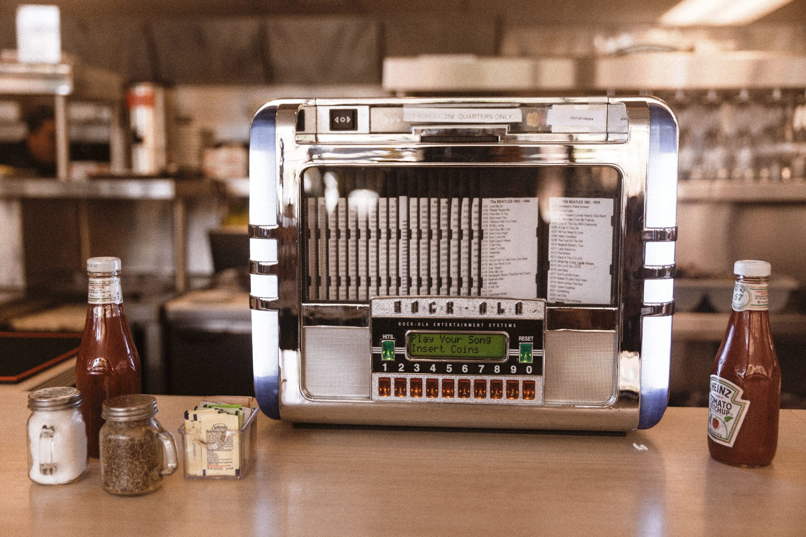 A vintage tabletop jukebox sits on a counter in a diner, flanked by salt and pepper shakers, sugar packets, and bottles of ketchup. The background shows stainless steel surfaces and diner equipment.