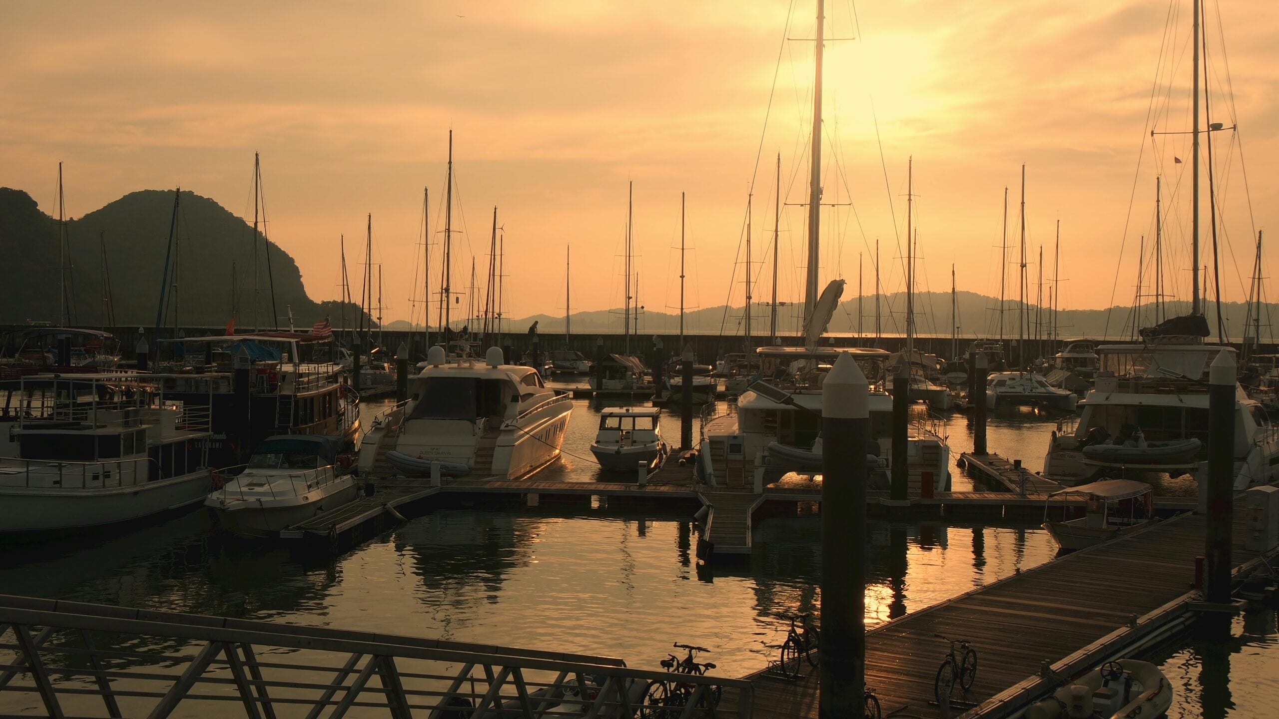 A marina at sunset with numerous boats and yachts docked, tall sailboat masts silhouetted against an orange sky, and calm water reflecting the warm light. Hills are visible in the background.