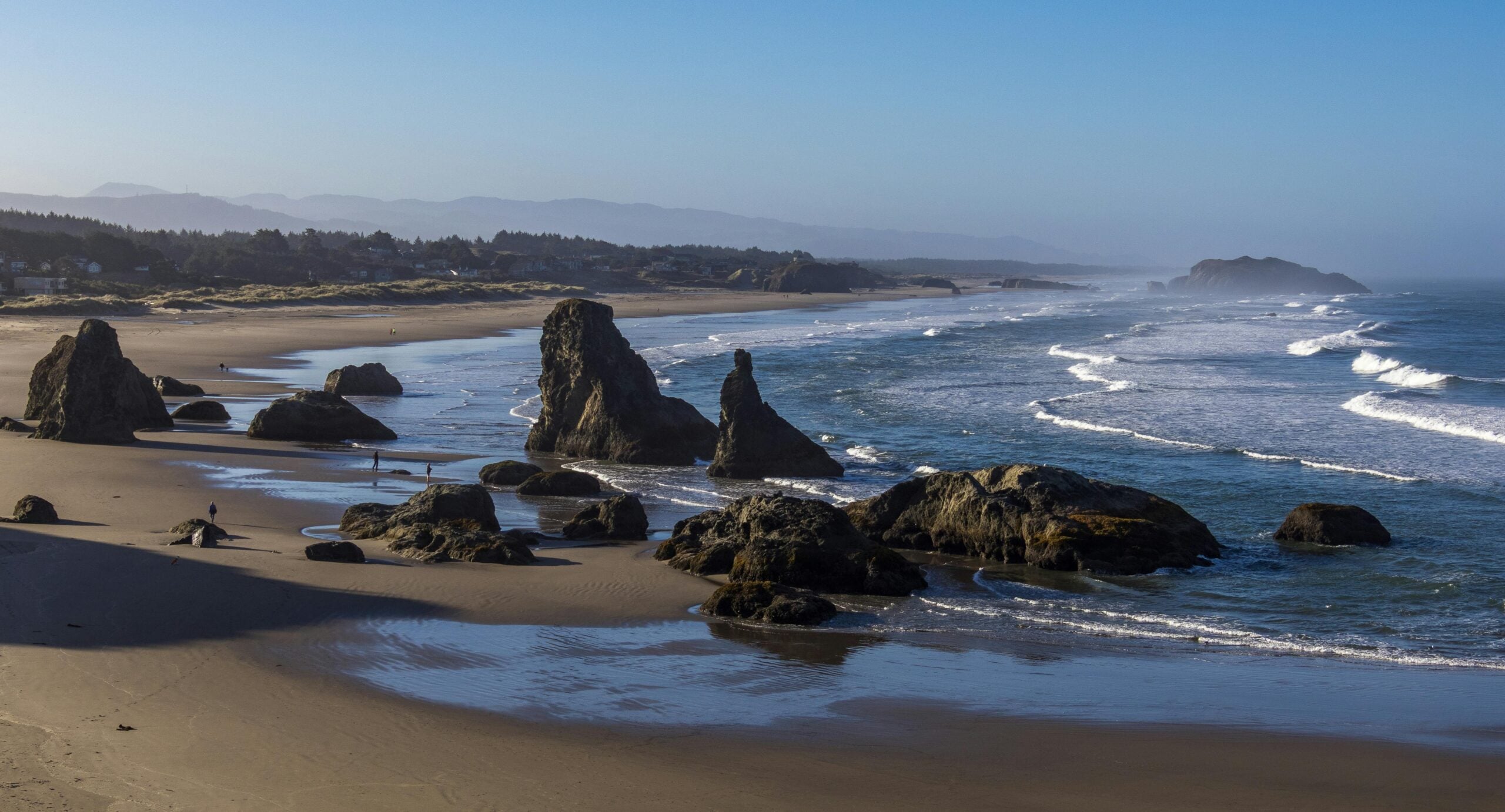 Large sea stacks and rocks dot a sandy beach along a rugged coastline with gentle waves and distant mountains under a clear blue sky. A few people walk along the shore.