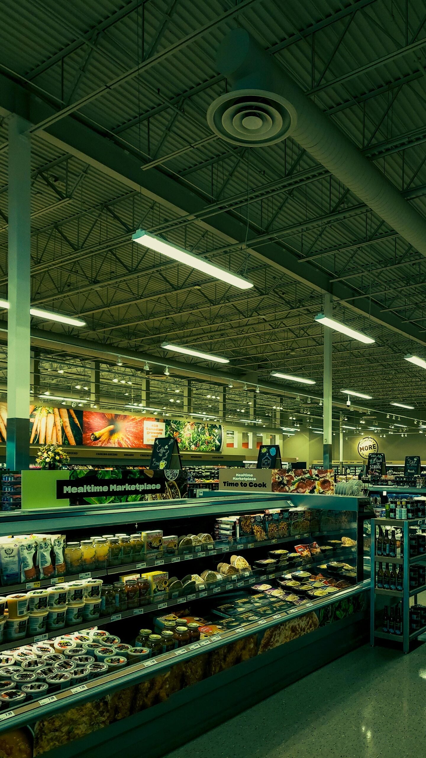 Wide view of a grocery store interior with refrigerated shelves stocked with prepared foods, dairy, and drinks; bright overhead lighting and colorful produce displays in the background.
