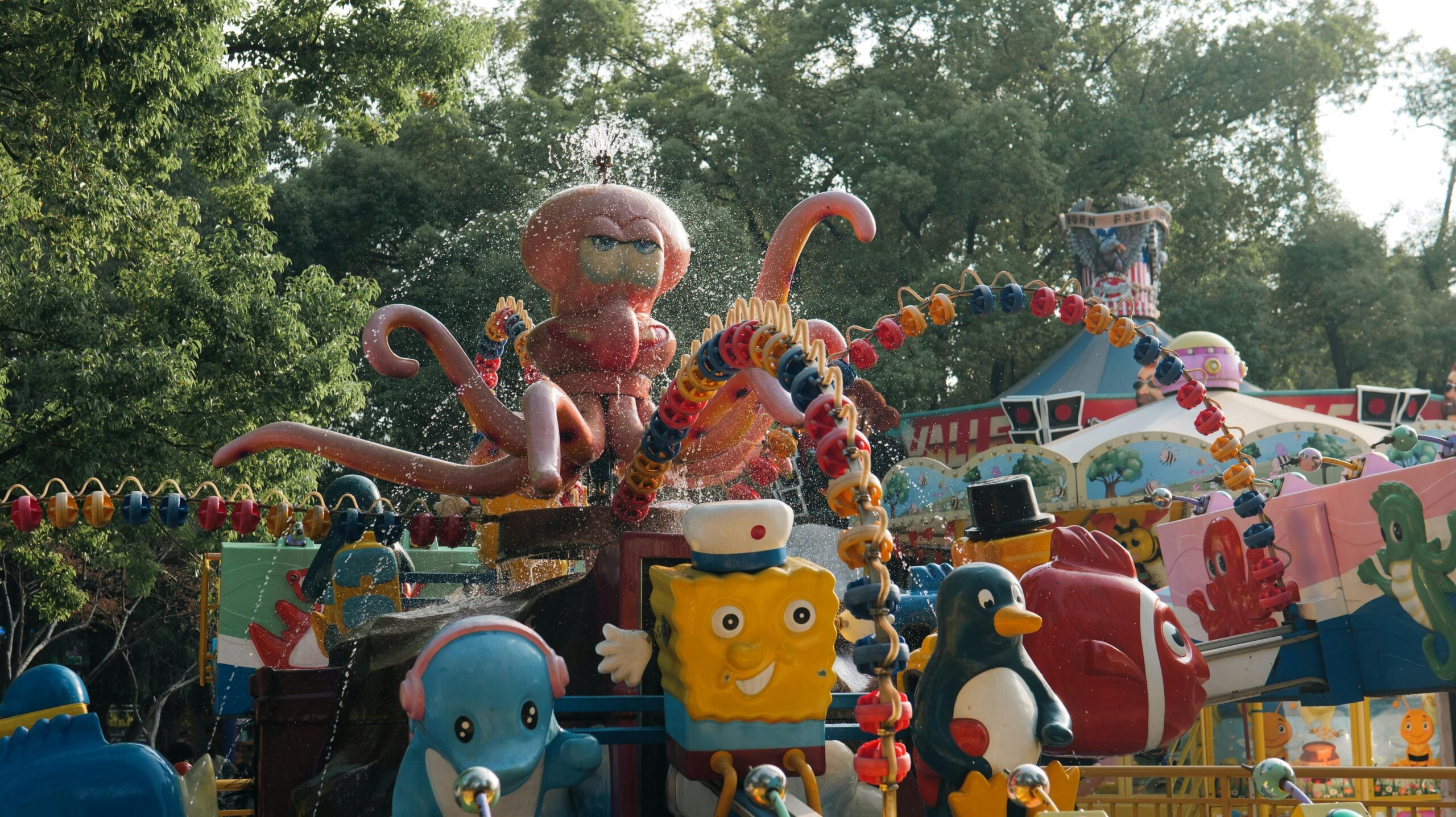 A colorful amusement park ride features cartoon characters, including a yellow sponge and a blue penguin, with a large pink octopus spraying water in the background. Trees and other ride structures are visible nearby.