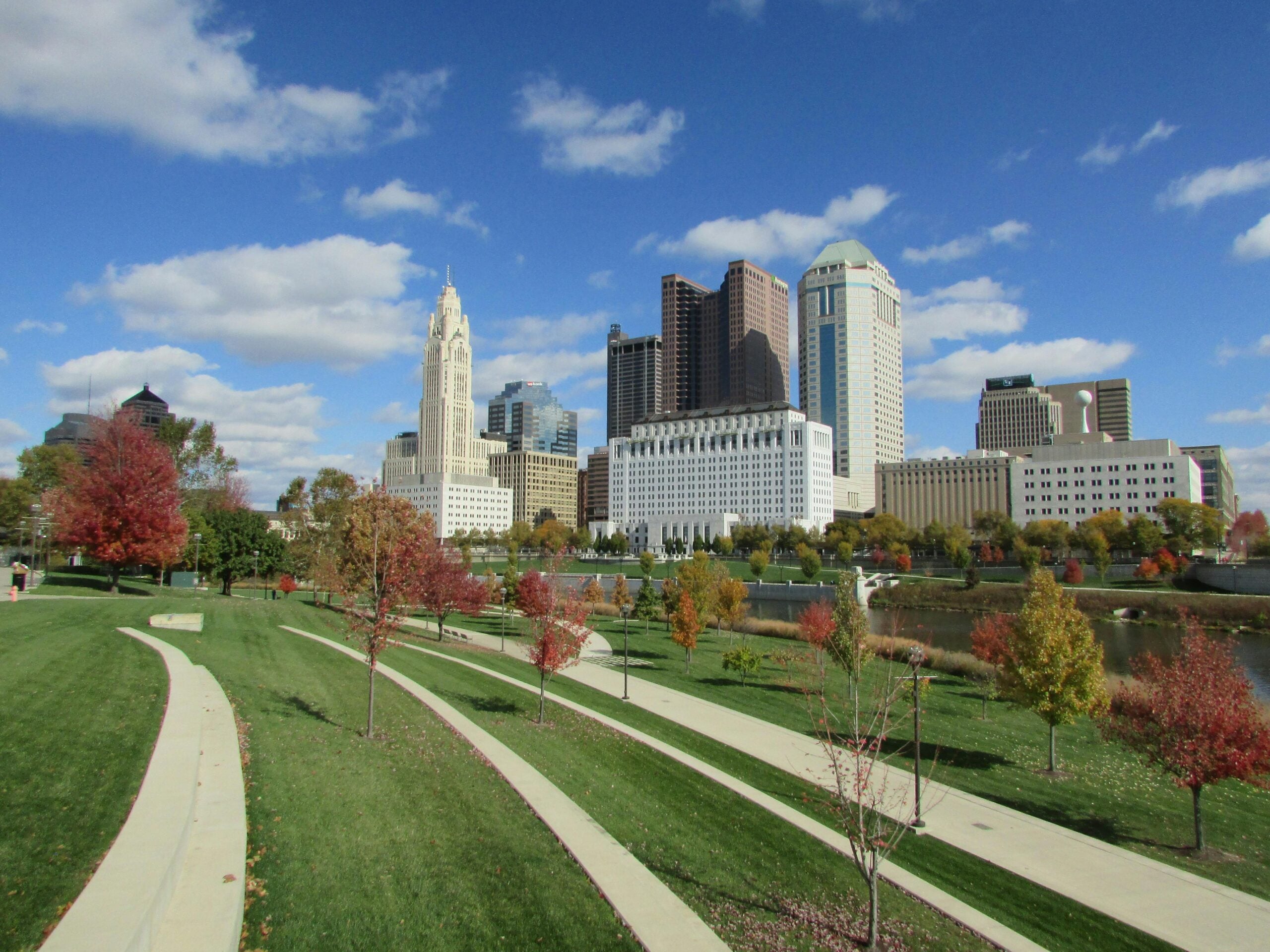 A city skyline with tall buildings rises behind a park featuring winding paths, green grass, and colorful autumn trees under a bright blue sky with scattered clouds.