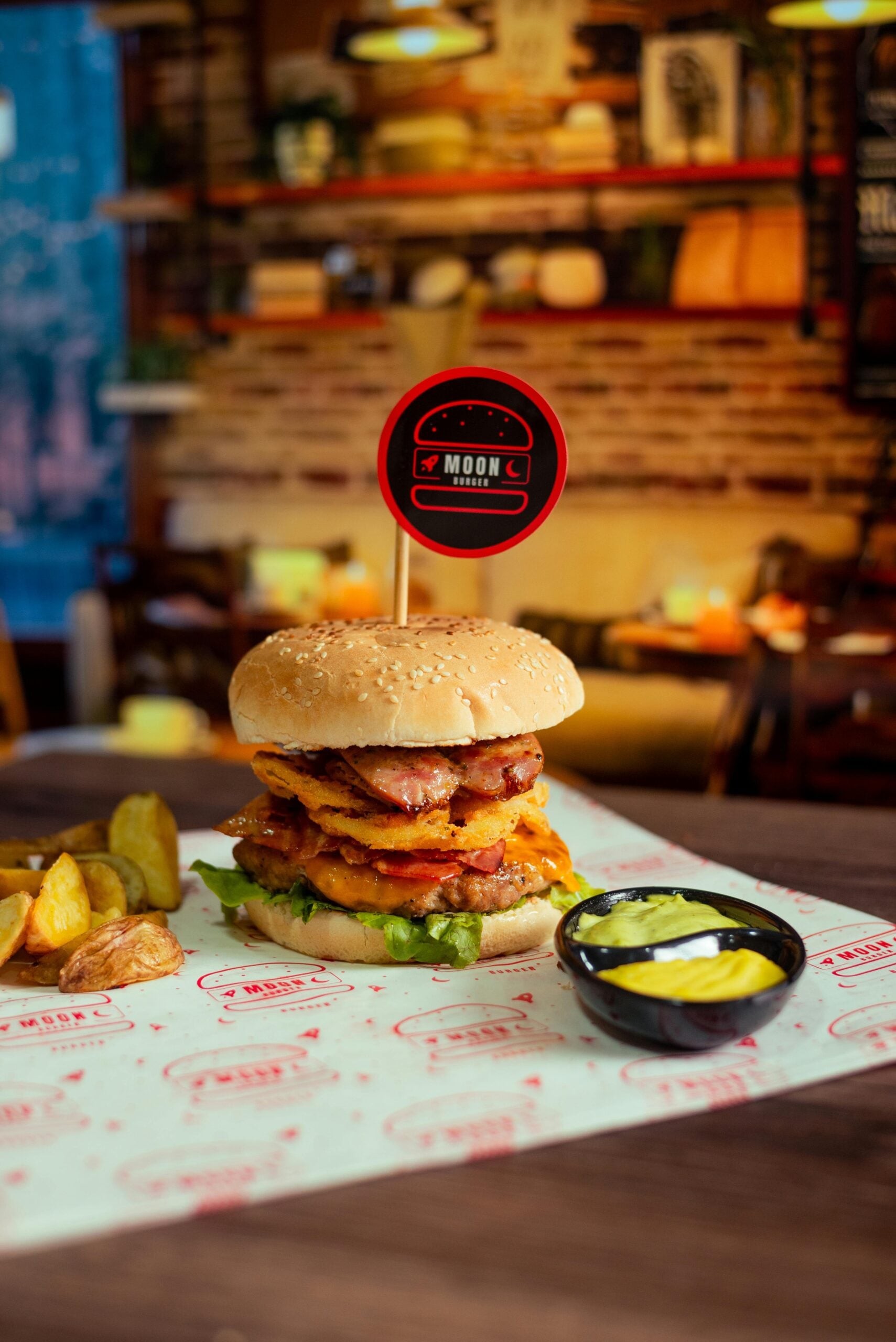 A gourmet burger with lettuce, tomato, onion rings, cheese, and sauce sits on a table with potato wedges and a small cup of yellow dip; a restaurant interior is blurred in the background.