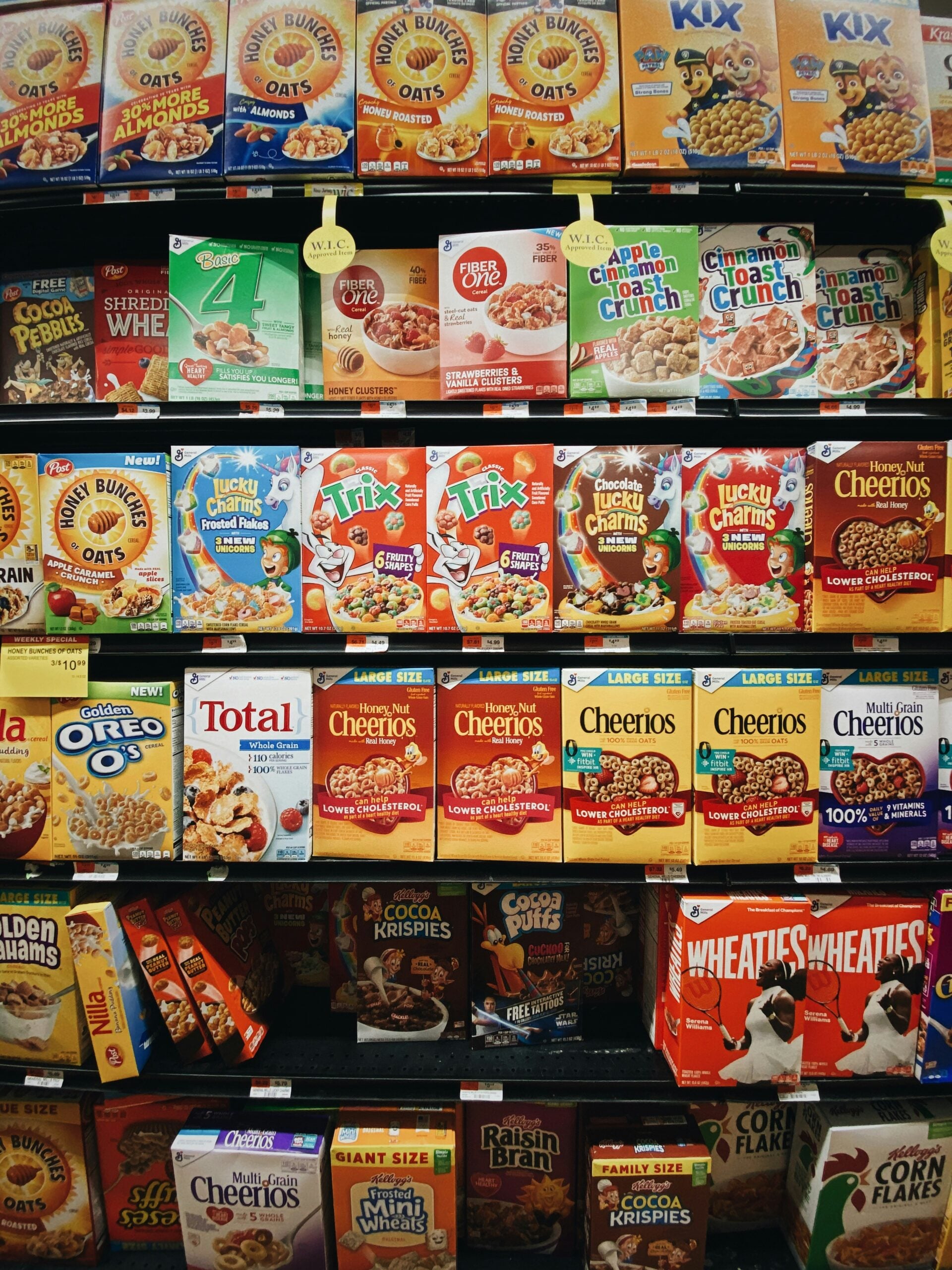 A grocery store shelf stocked with various colorful breakfast cereal boxes, including Cheerios, Trix, Lucky Charms, Cinnamon Toast Crunch, and Raisin Bran, arranged in multiple rows.