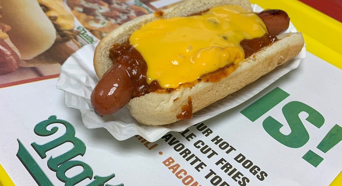 A classic fast-food hot dog in a bun, topped with chili and melted cheddar cheese, sits on a paper tray atop a yellow tray with part of a Nathan's Famous advertisement visible underneath.