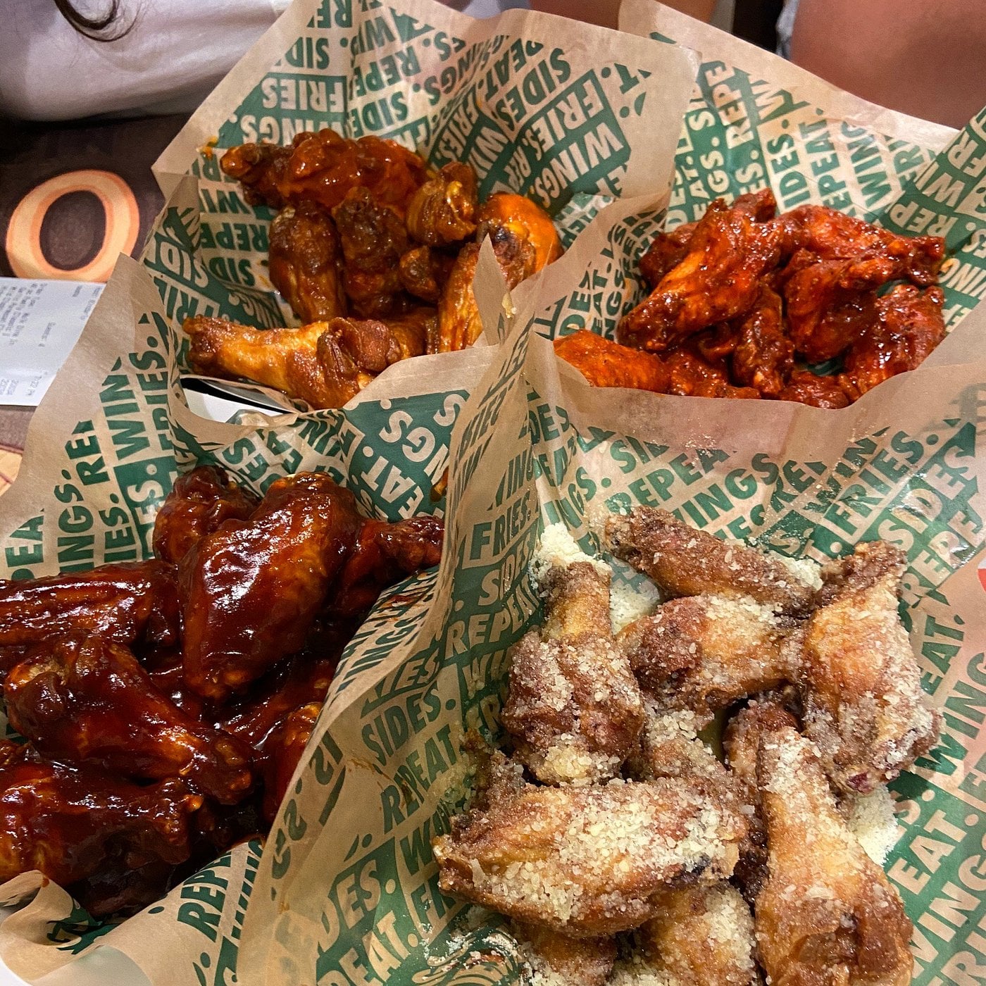 Four baskets of chicken wings with different seasonings and sauces, including barbecue, garlic parmesan, and spicy varieties, served on green and brown Wingstop paper.