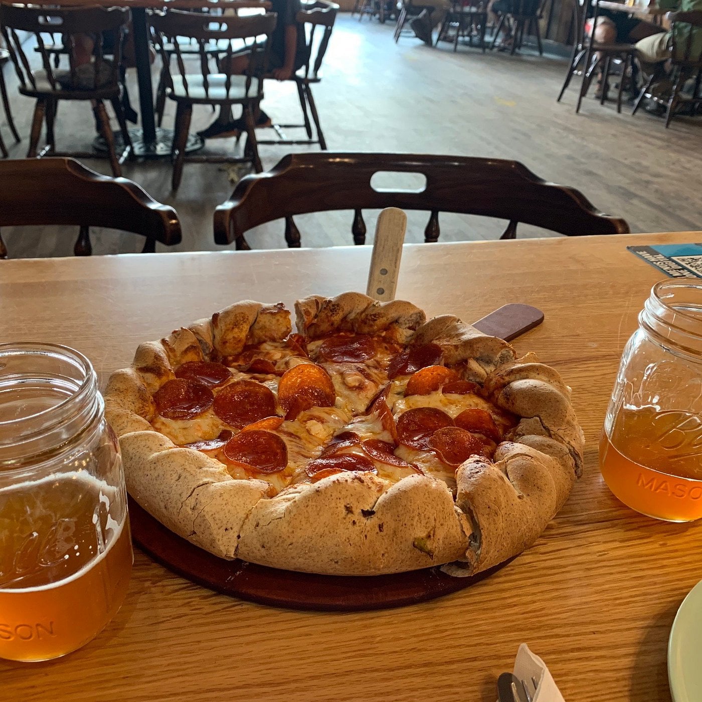 A pepperoni pizza with a thick crust and a knife in the center sits on a wooden table, flanked by two mason jars of light-colored beverages—perfect for road trip food stops in a rustic restaurant setting.