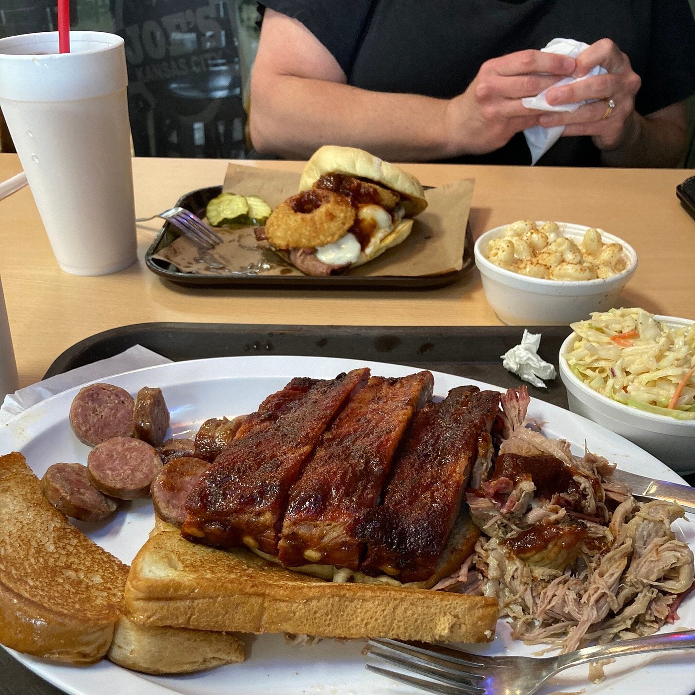 A tray loaded with ribs, sliced sausage, pulled pork, Texas toast, coleslaw, and a drink sits ready—classic road trip food stops fare. Nearby, someone grabs a napkin near a sandwich with pickles and creamy macaroni and cheese.