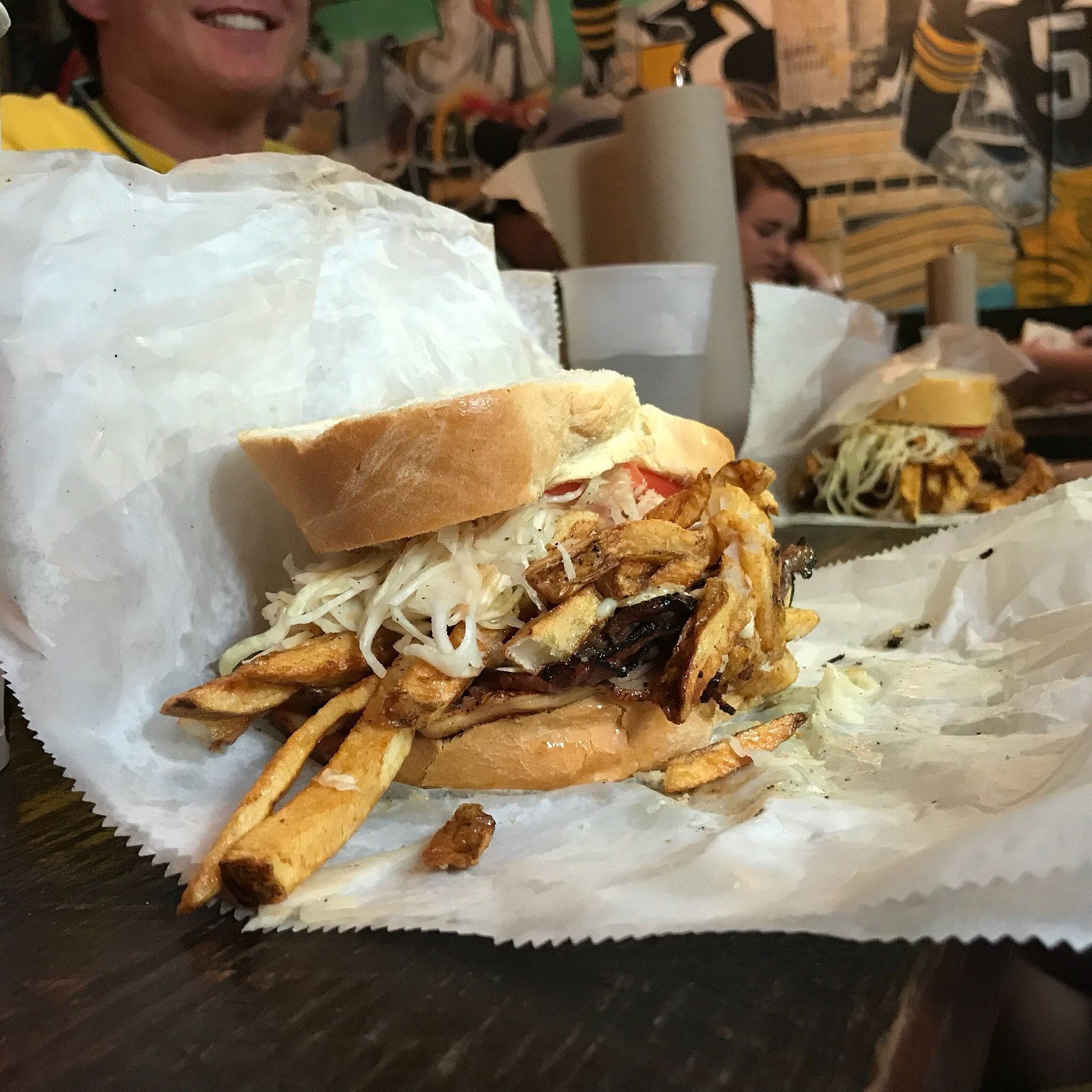 A close-up of a sandwich filled with fries, coleslaw, and meat on wax paper, with people and colorful wall art in the background at a casual restaurant—a perfect find for road trip food stops.