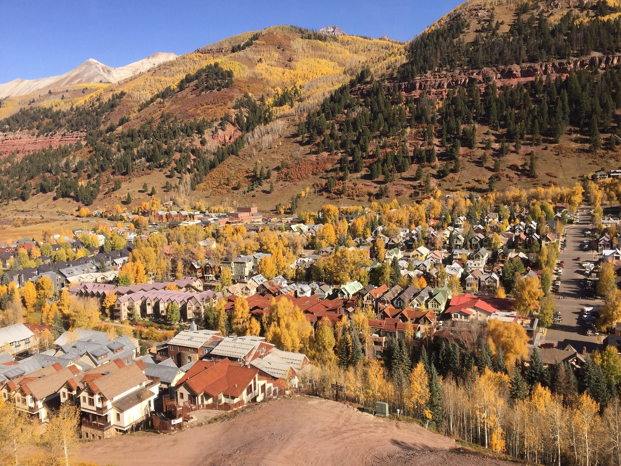 Aerial view of a unique American town nestled in a valley, with rows of houses surrounded by yellow autumn trees and forested hills in the background under a clear blue sky.