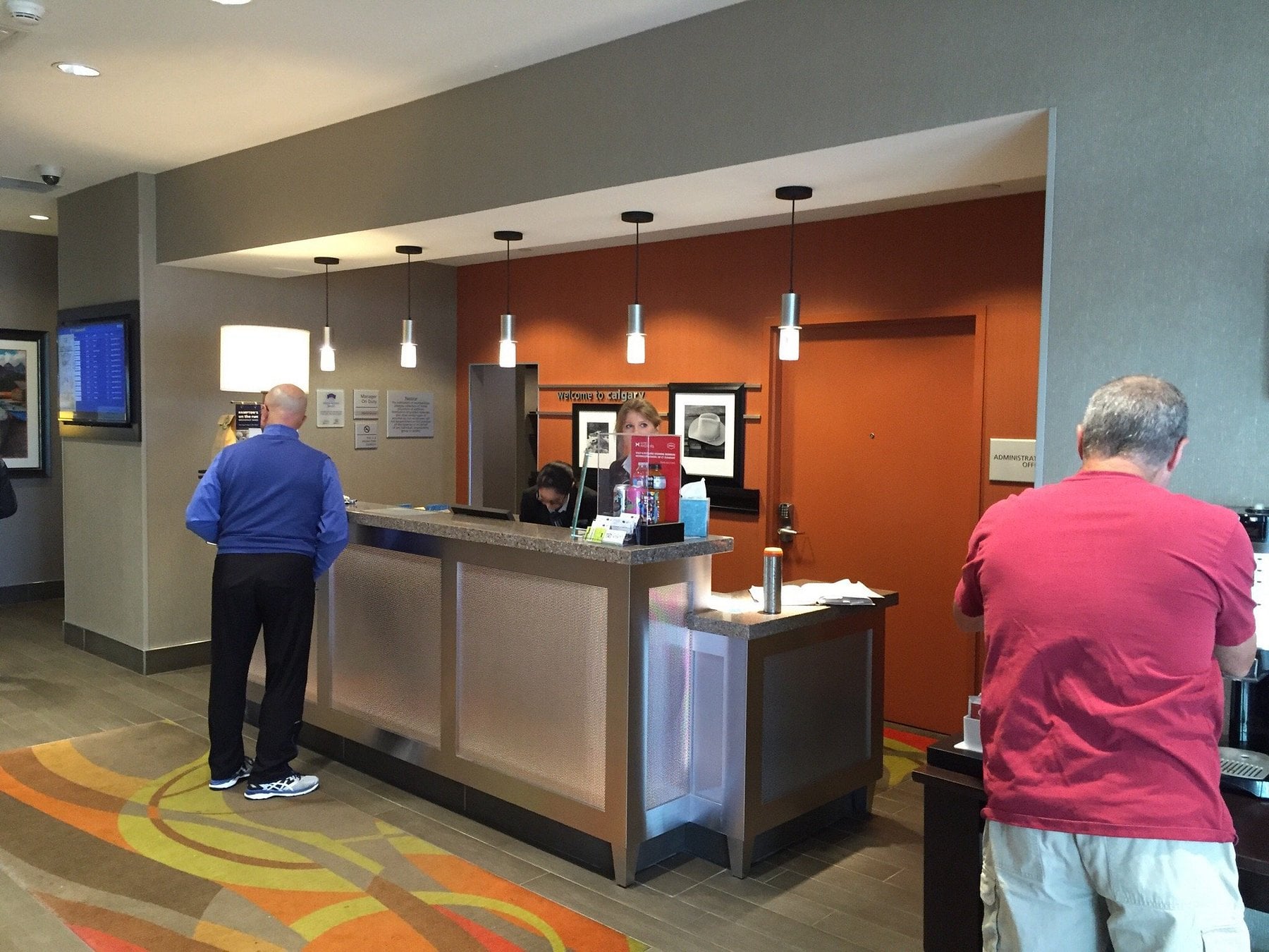 A hotel front desk area with a staff member assisting a guest. Another man in a blue jacket stands at the counter, while a man in a red shirt uses a coffee station nearby. The modern lobby reflects Hotel Brands 2026 style with warm lighting and décor.