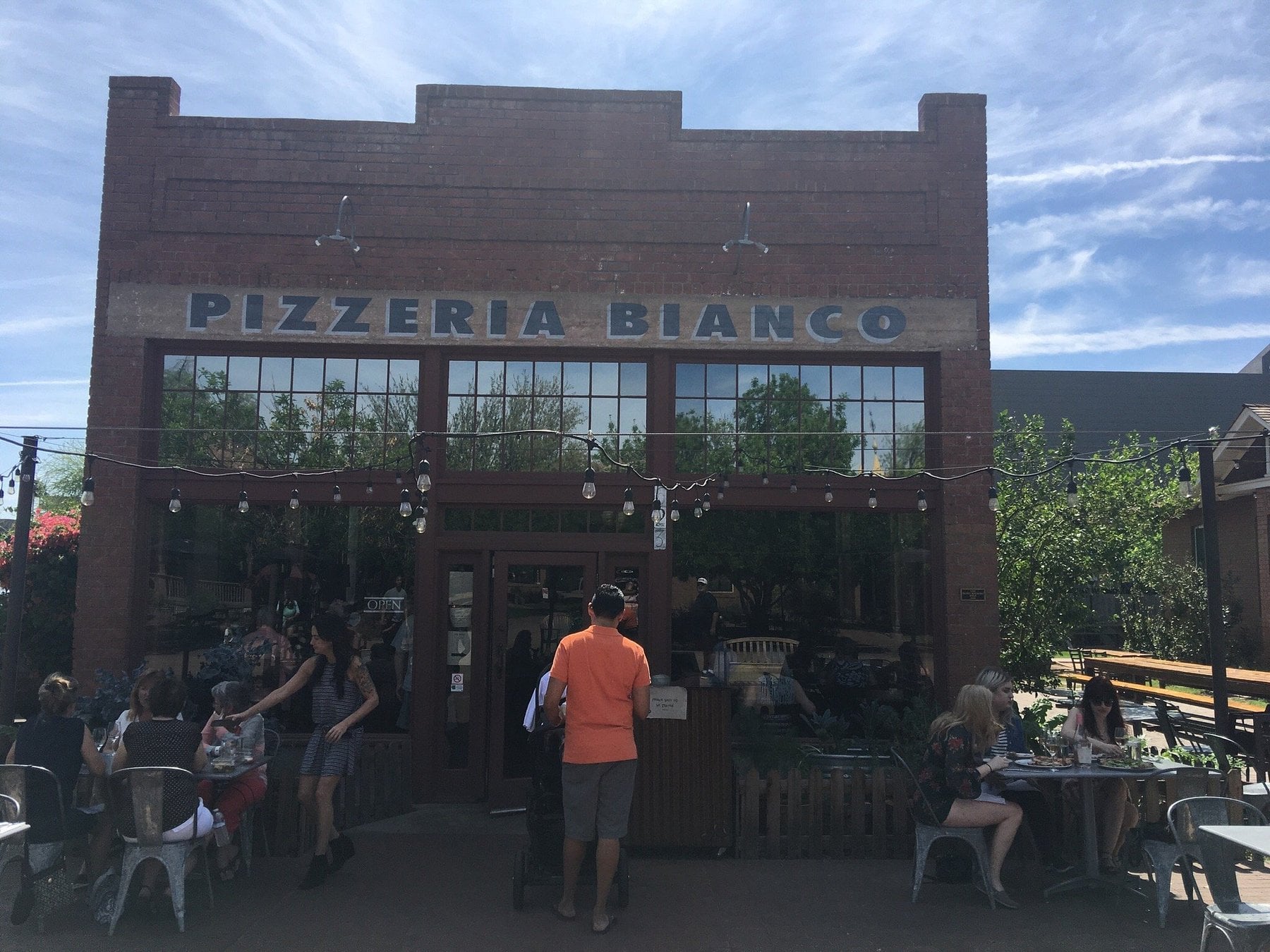 A brick building with large windows and a sign reading "Pizzeria Bianco." Popular as one of the best road trip food stops, people are dining at outdoor tables under string lights on a sunny day.