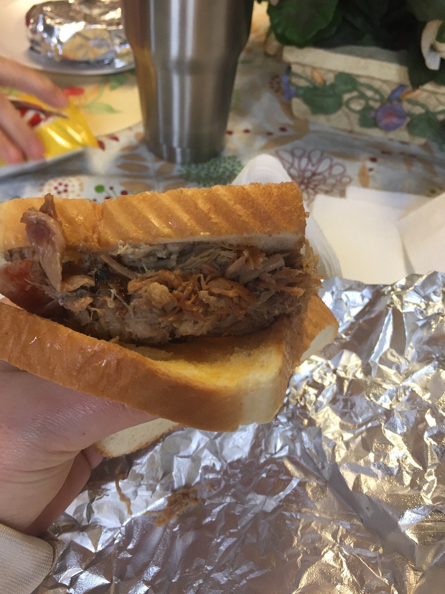 A hand holds a sandwich made with pulled pork between two slices of white bread, resting on aluminum foil—classic road trip food stops in mind. In the background, a metal tumbler and plate sit among other items on the table.