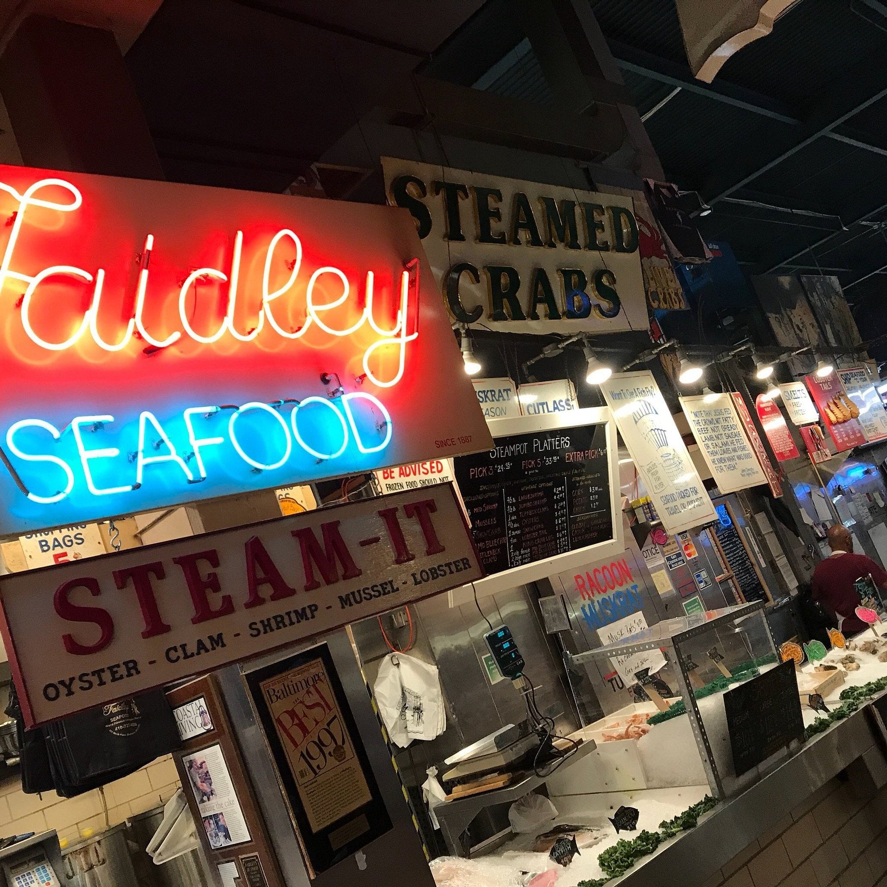 A bustling indoor seafood market, perfect for road trip food stops, with neon signs reading “Faidley Seafood” and “STEAMED CRABS.” Counters display fresh seafood as menu boards and various signs hang above.