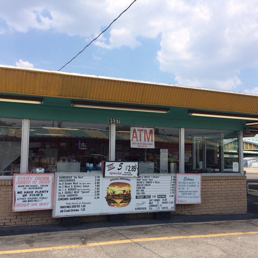 A small roadside diner with yellow trim displays menus and signs in its front windows, including burger specials and an ATM sign above the counter—a classic example of the charm found in roadside restaurants. The sky is partly cloudy.