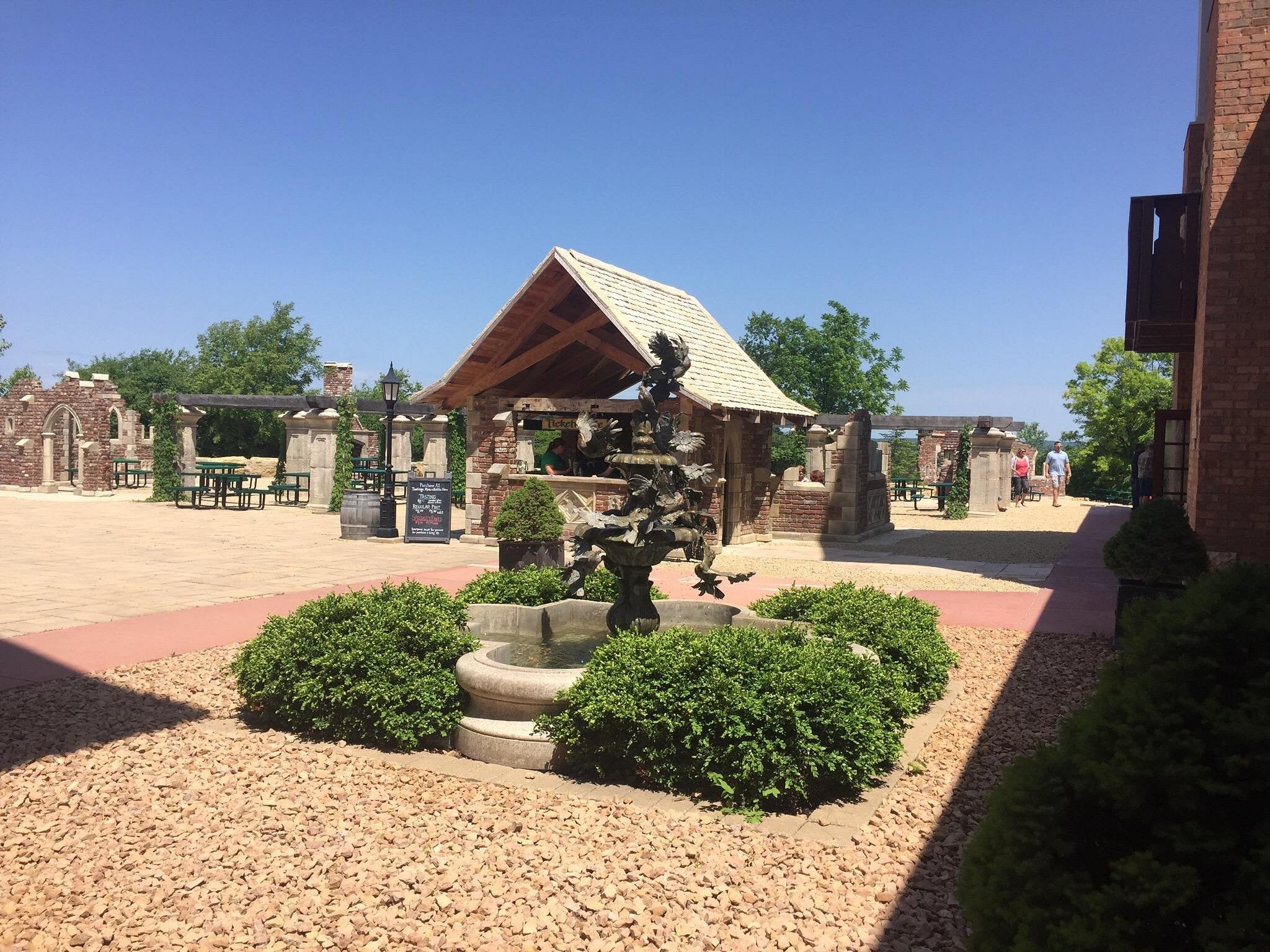 A courtyard with a central fountain surrounded by greenery, stone paths, picnic tables under a wooden pavilion, and brick and stone structures—capturing the charm found in Unique American Towns—rests under a clear blue sky.