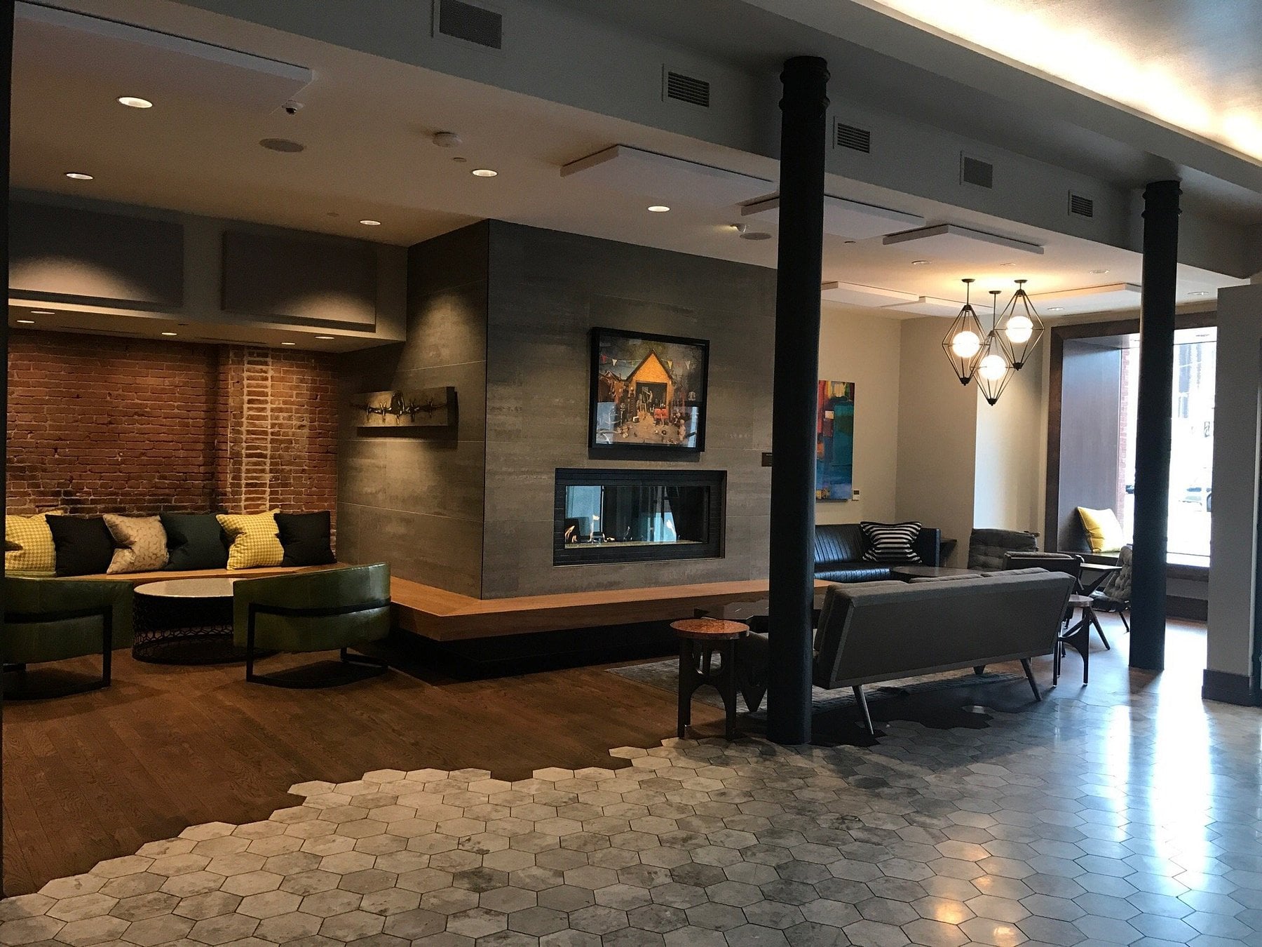A modern lounge in the style of Small-Town Luxury Hotels, featuring hexagonal tile flooring, wood floors, green chairs, sofas, a fireplace, brick walls, contemporary art, and floor-to-ceiling windows welcoming natural light.