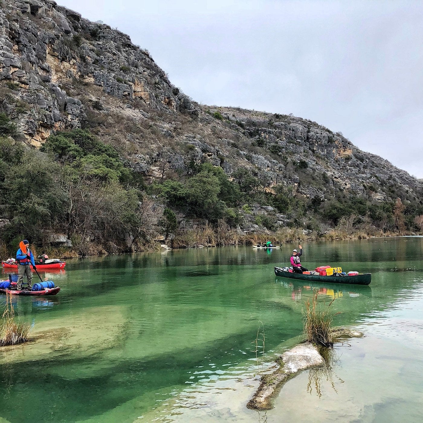Several people paddle kayaks on a clear green river bordered by rocky hills and sparse vegetation on a cloudy day—a peaceful scene found in one of the unique American towns known for its calm water and natural surroundings.