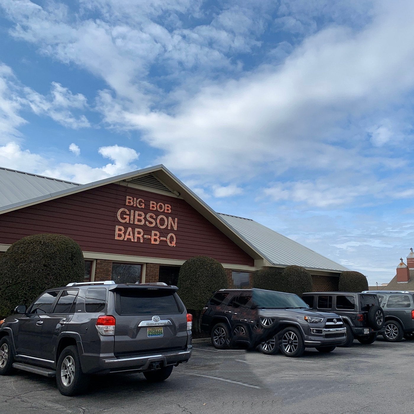 Four SUVs and a Jeep are parked outside Big Bob Gibson Bar-B-Q, a classic road trip food stop housed in a red and brown building with trimmed bushes in front, under a partly cloudy sky.
