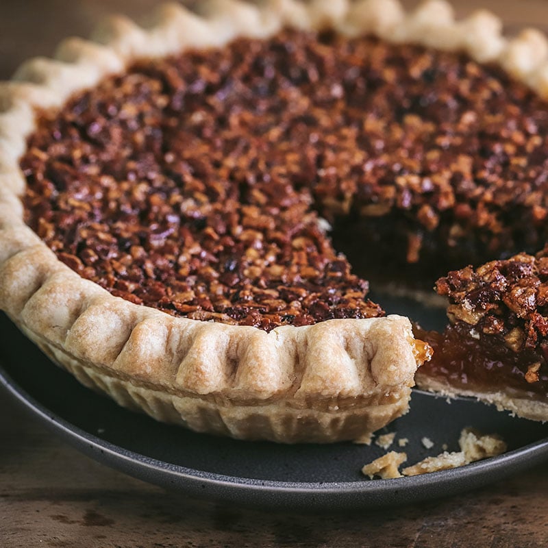 A pecan pie with a golden, flaky crust sits on a dark plate. A slice has been cut and is being lifted out, revealing the nutty, caramel filling inside.