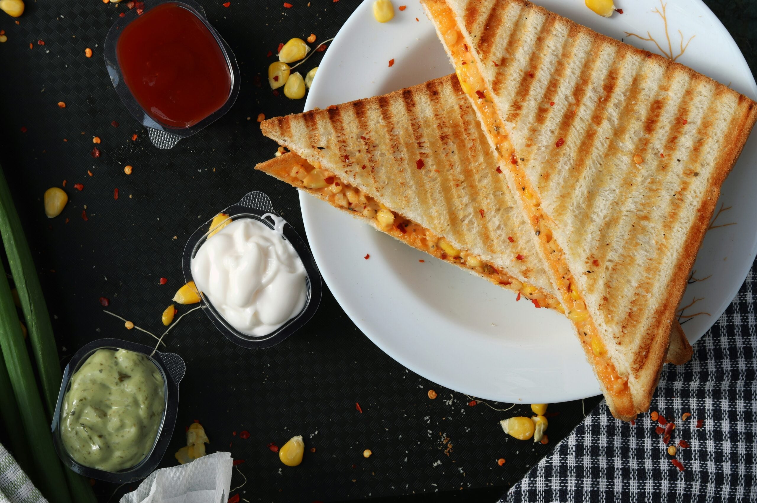 Two grilled sandwich triangles are served on a white plate, with small containers of ketchup, mayonnaise, and green sauce nearby. Some corn kernels and a checkered cloth are also visible on a dark surface.