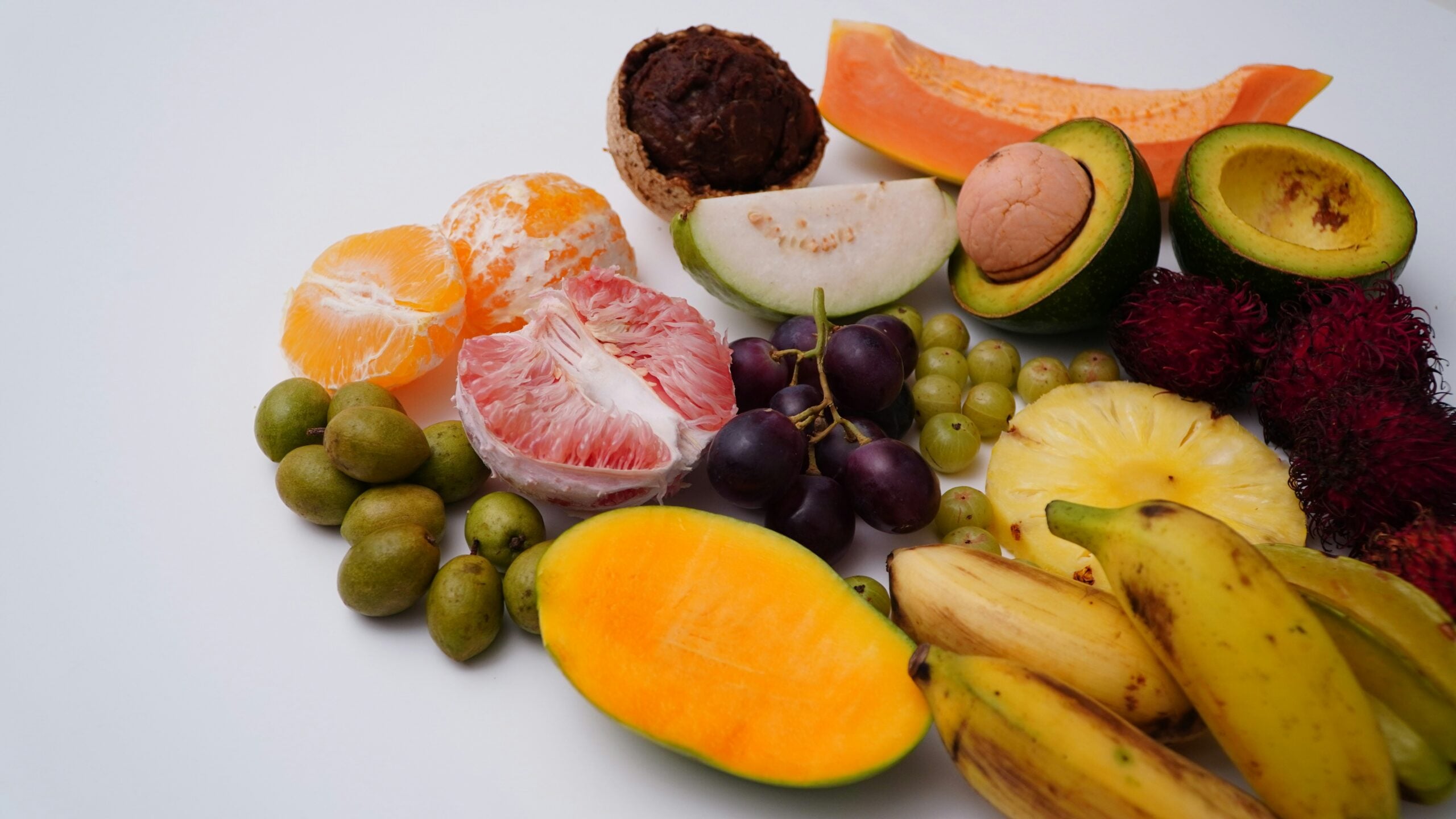 A colorful assortment of tropical fruits, including bananas, mango, grapes, pineapple, guava, orange, pomelo, avocado, papaya, rambutan, and sapote, arranged on a white background.