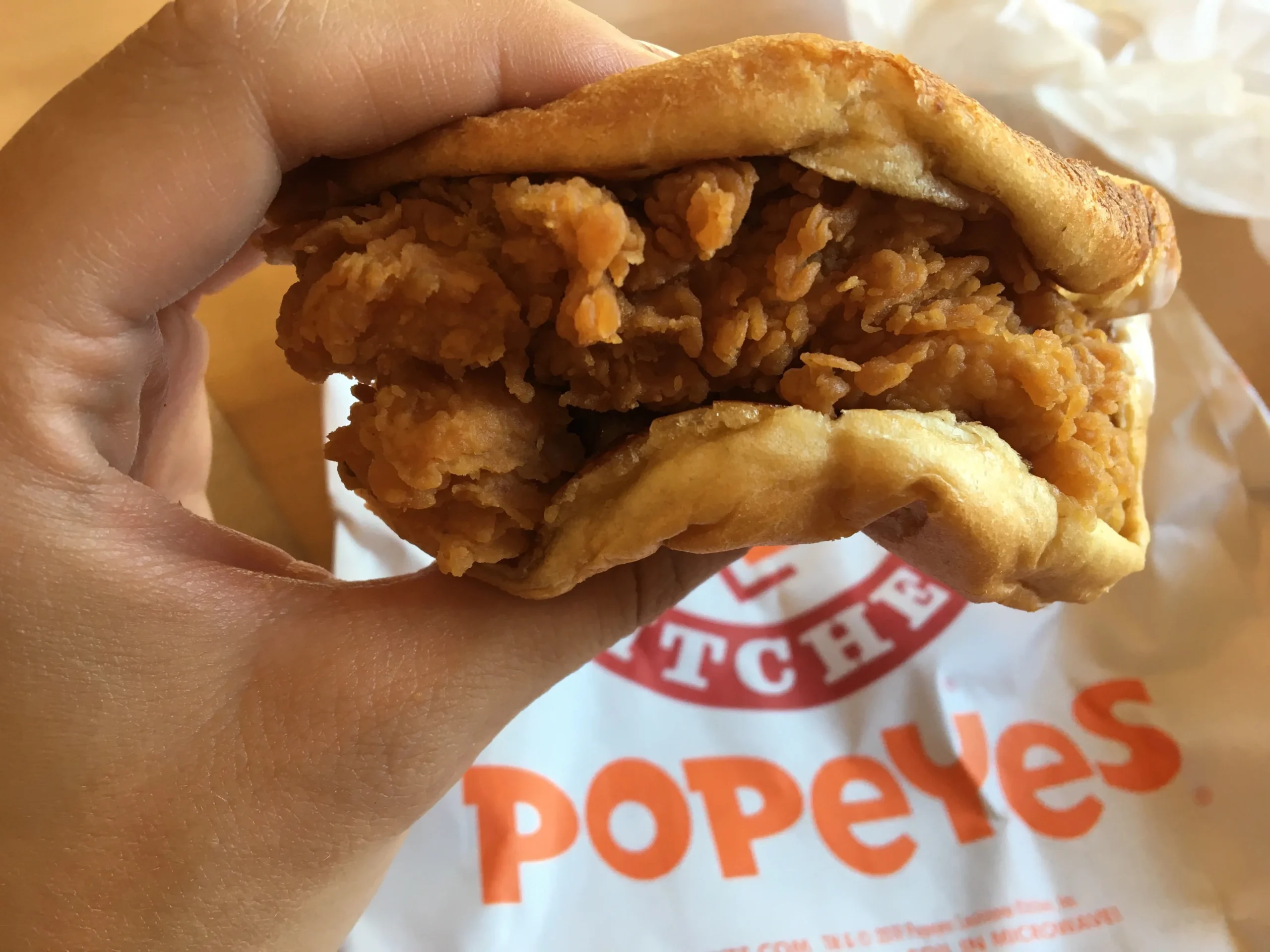 A hand holds a crispy fried chicken sandwich in front of a Popeyes wrapper, showing the sandwich's golden, crunchy chicken fillet inside a soft bun.