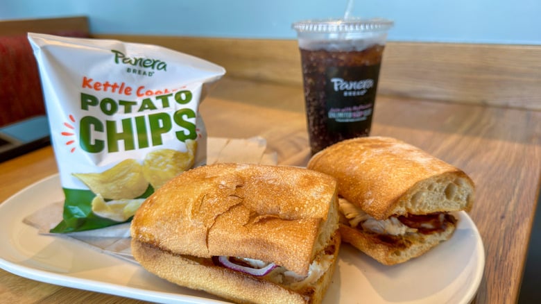 A plate with two halves of a sandwich on ciabatta bread sits next to a bag of Panera kettle-cooked potato chips and a cup of dark soda with a straw on a table in a Panera Bread restaurant.