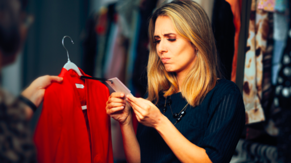 A woman with blonde hair looks closely at the price tag of a red shirt on a hanger, appearing uncertain or puzzled&mdash;perhaps contemplating delayed purchases&mdash;while standing in a clothing store with racks of clothes in the background.