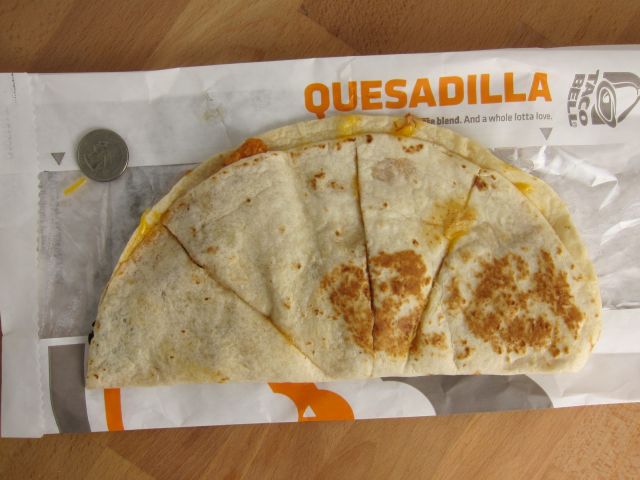 A Taco Bell quesadilla cut into four pieces sits on its wrapper next to a quarter on a wooden surface. The word "QUESADILLA" is visible on the wrapper.