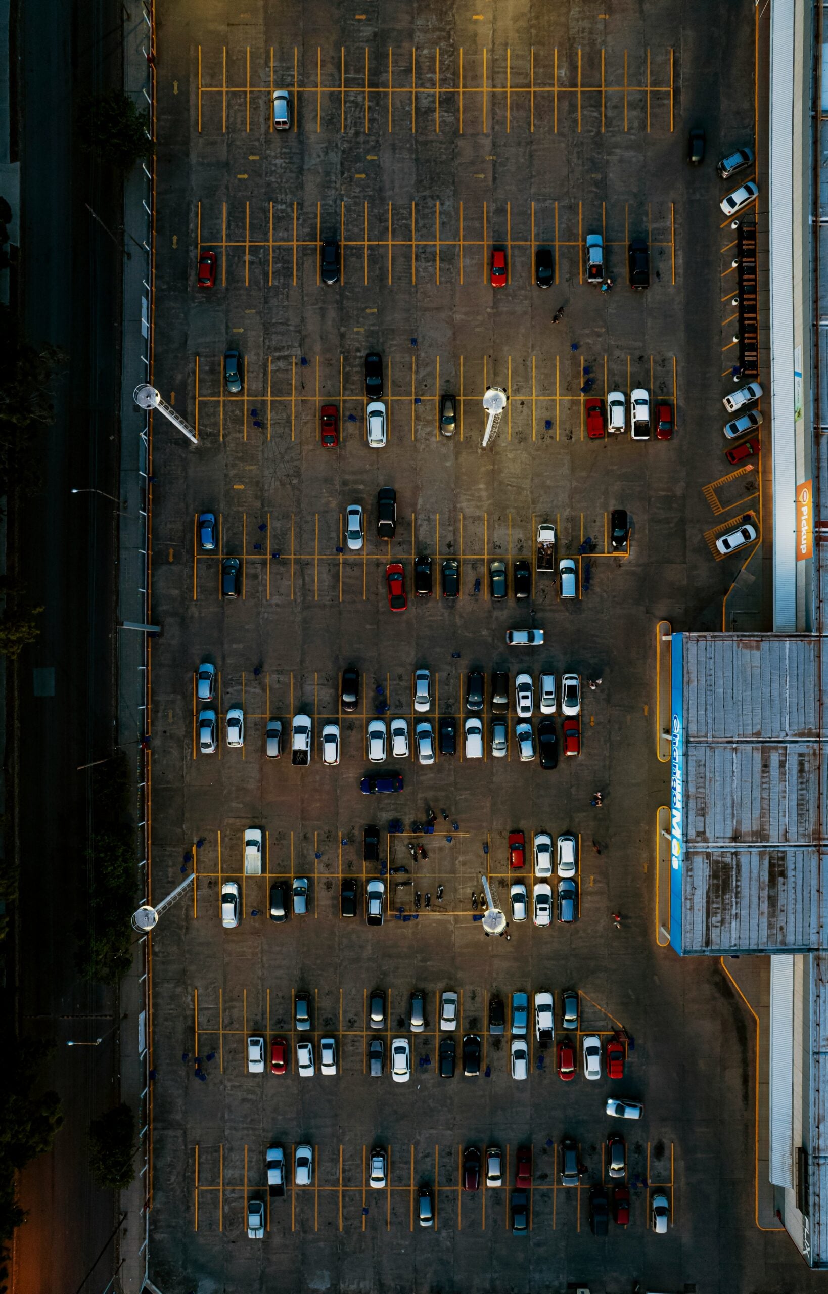 Aerial view of a large parking lot with many cars parked in rows, mostly white and red vehicles, with some empty parking spaces. The lot is next to a building and illuminated by tall streetlights.