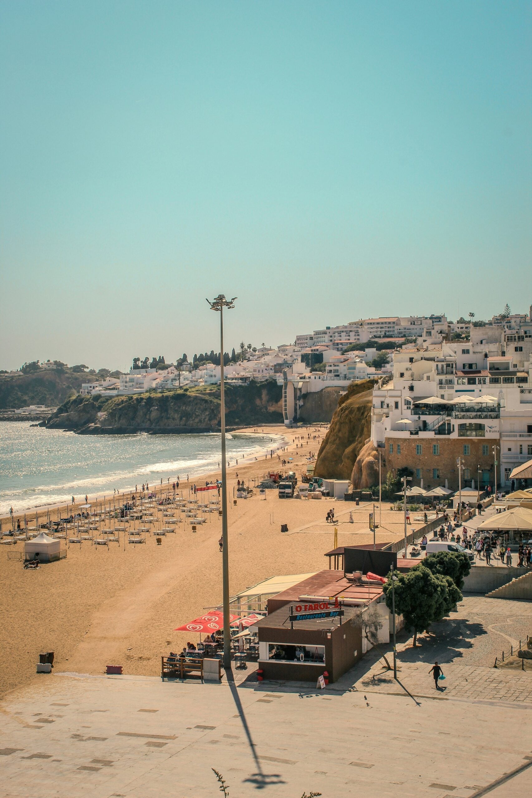 A sunny beach with light golden sand, scattered umbrellas, and people walking. White buildings line the hillside overlooking the shoreline, and the calm sea stretches under a clear blue sky.