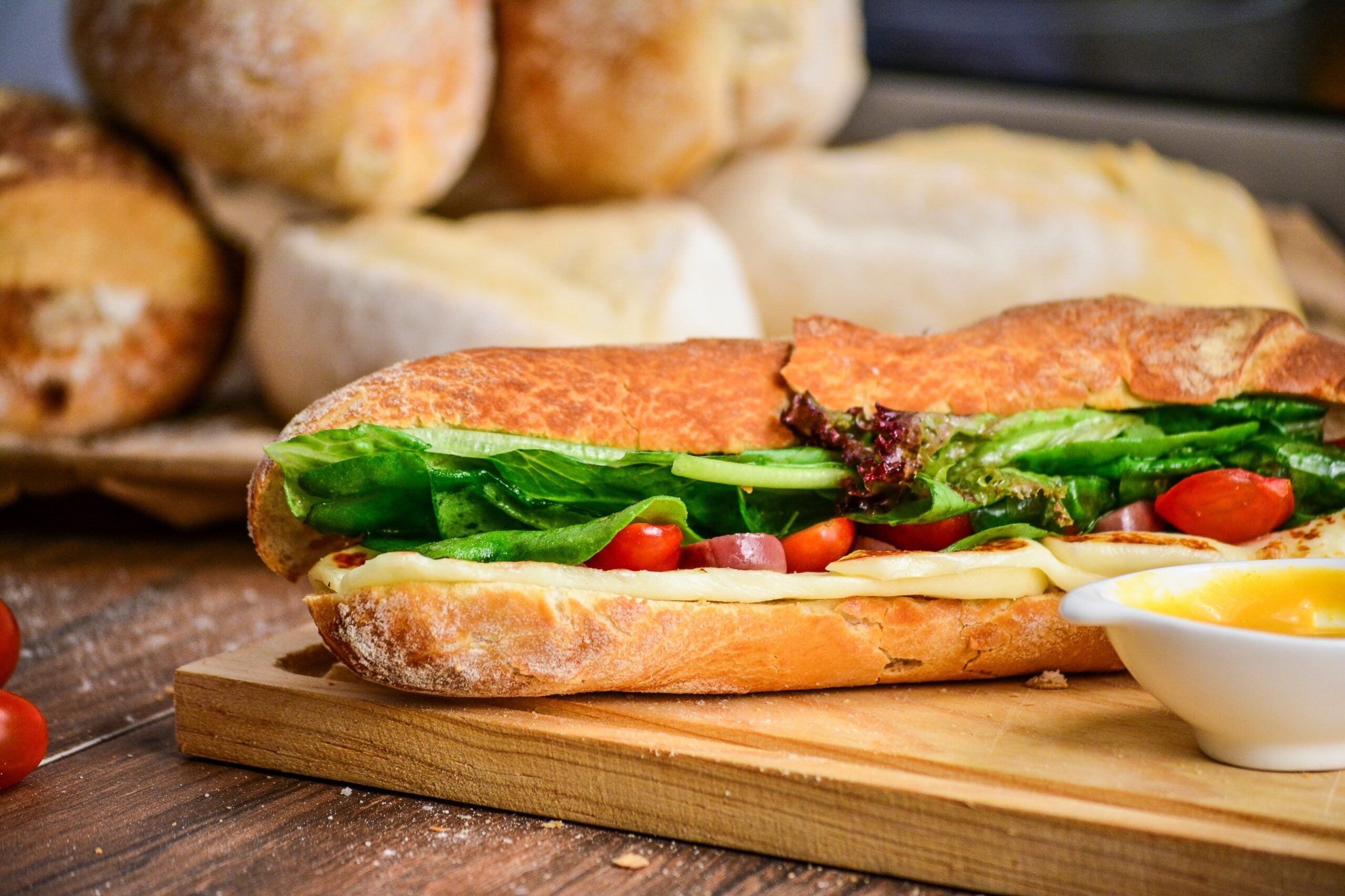 A fresh sandwich with lettuce, tomato, cheese, and possibly ham in a crusty baguette sits on a wooden board next to a small dish of yellow sauce. Loaves of bread are visible in the background.