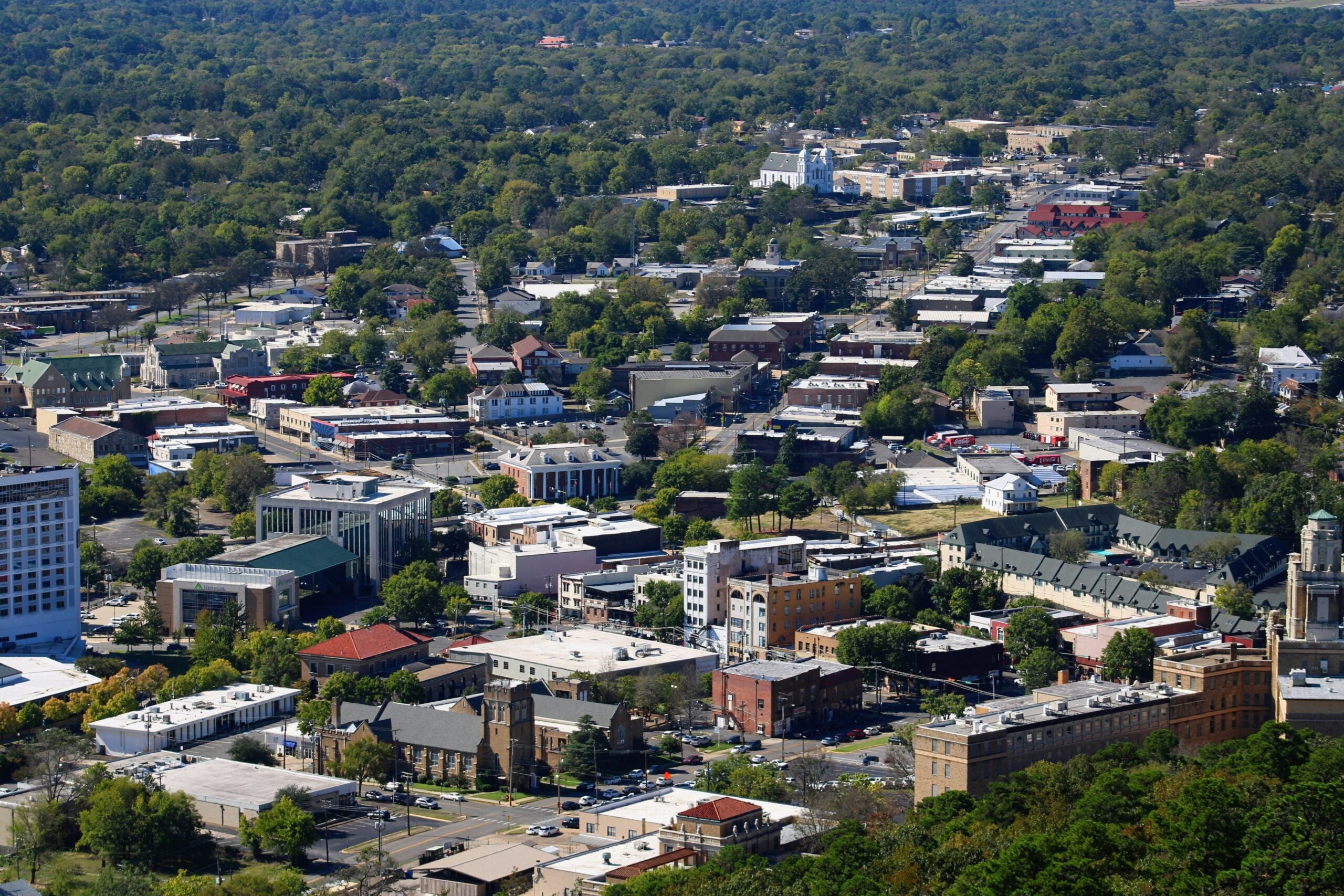 Aerial view of a small city with tree-lined streets, numerous low-rise buildings, and surrounding green forests on a sunny day.