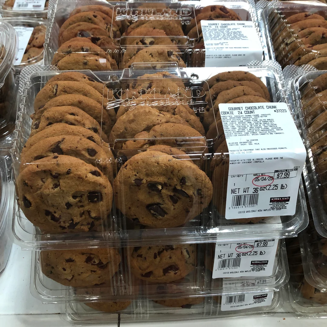 Plastic containers filled with chocolate chip cookies are stacked on a store shelf. Each container has a white label with product and pricing information. The cookies are golden brown with visible chocolate chunks.