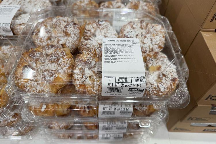 Clear plastic containers filled with powdered sugar-topped pastries are stacked on a store shelf. A label on the top container displays product details, weight, and price. Cardboard boxes are stacked nearby.
