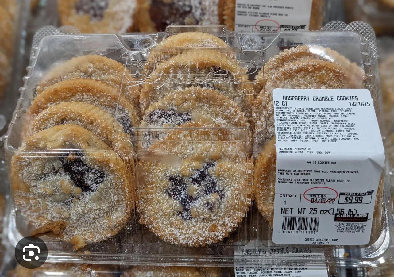 A clear plastic container holds twelve raspberry crumble cookies dusted with powdered sugar. The label on the lid details the product name, ingredients, nutrition facts, and price.