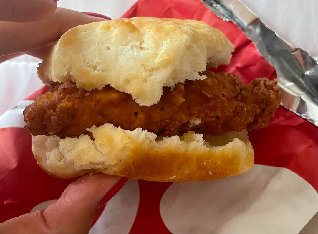A hand holds a chicken biscuit sandwich with a piece of crispy fried chicken between two halves of a fluffy biscuit, against a red and white wrapper background.