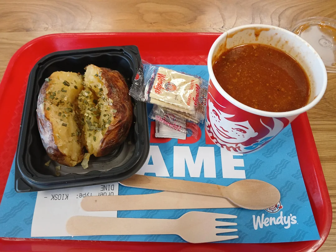 A Wendy’s tray with a baked potato topped with chives in a black container, a cup of chili, cracker packets, a wooden fork and spoon, and a Wendy’s placemat on a wooden table.