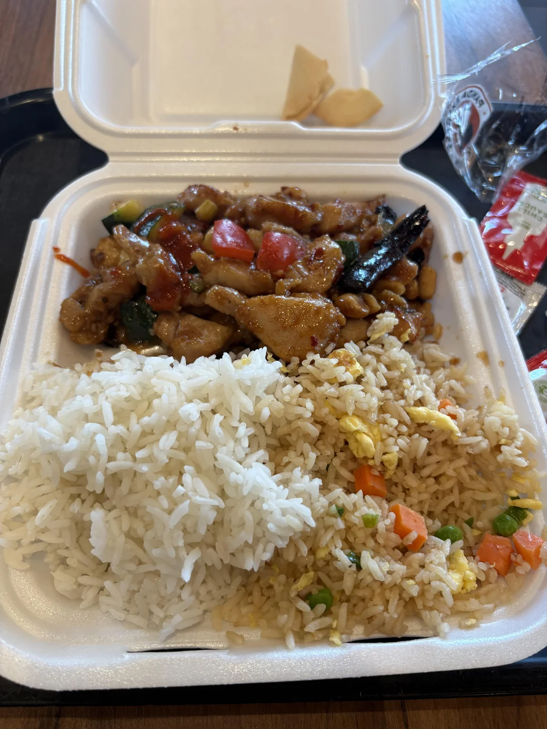 Fast-food secret menu hacks. A takeout container with white rice, fried rice with peas and carrots, and stir-fried chicken with vegetables in a brown sauce. Two pieces of crispy wonton are in the top section.