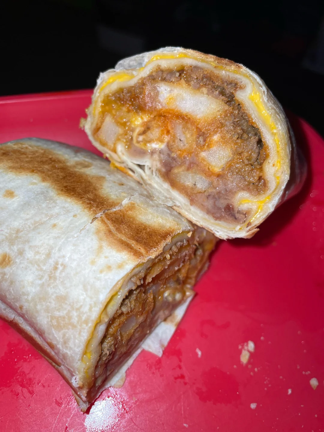 A close-up of a burrito cut in half on a red plate, showing layers of beans, cheese, sauce, and possibly potatoes or meat inside a toasted flour tortilla.